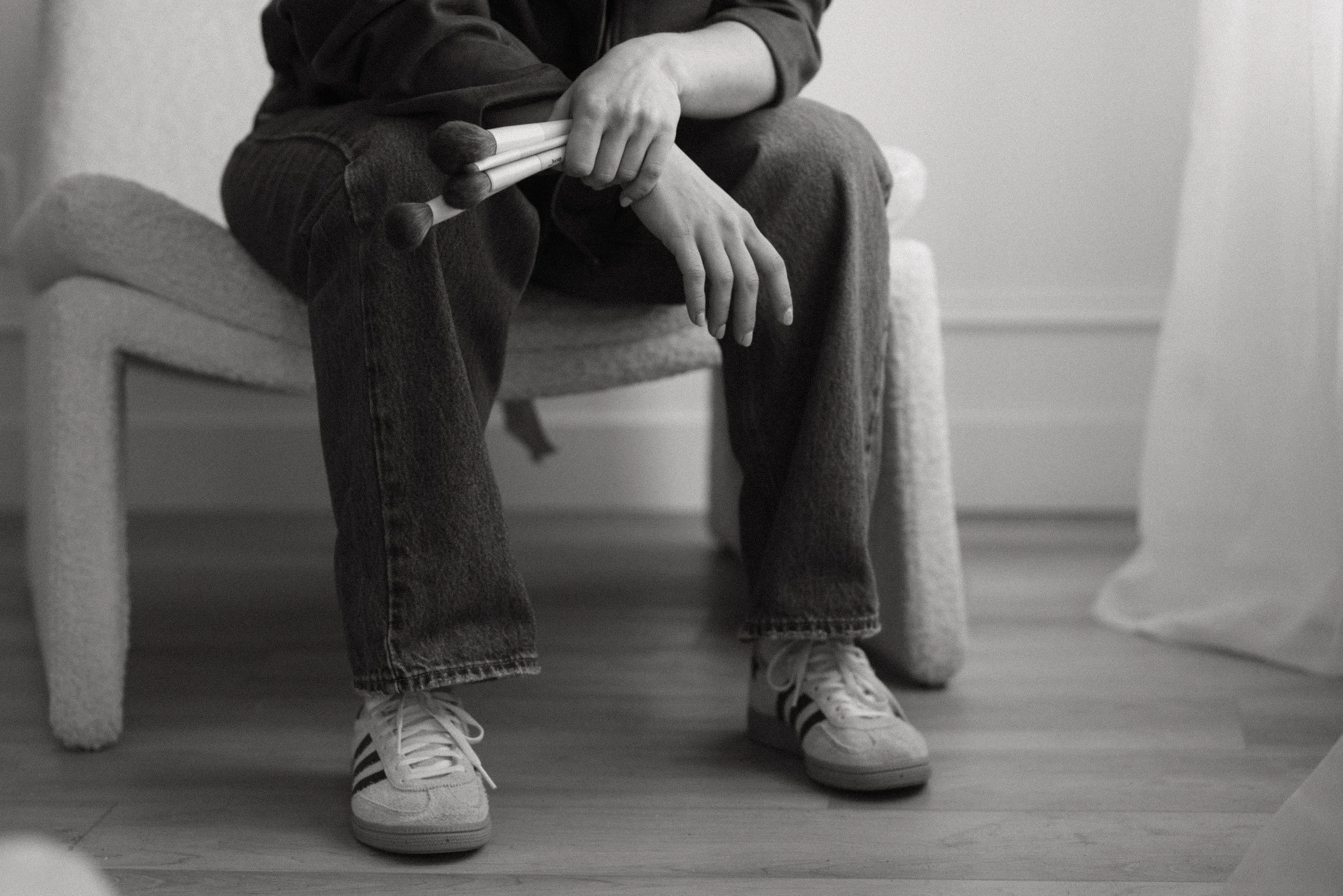 Person sitting on a chair holding a bunch of makeup brushes, wearing sneakers, jeans, and a jacket.