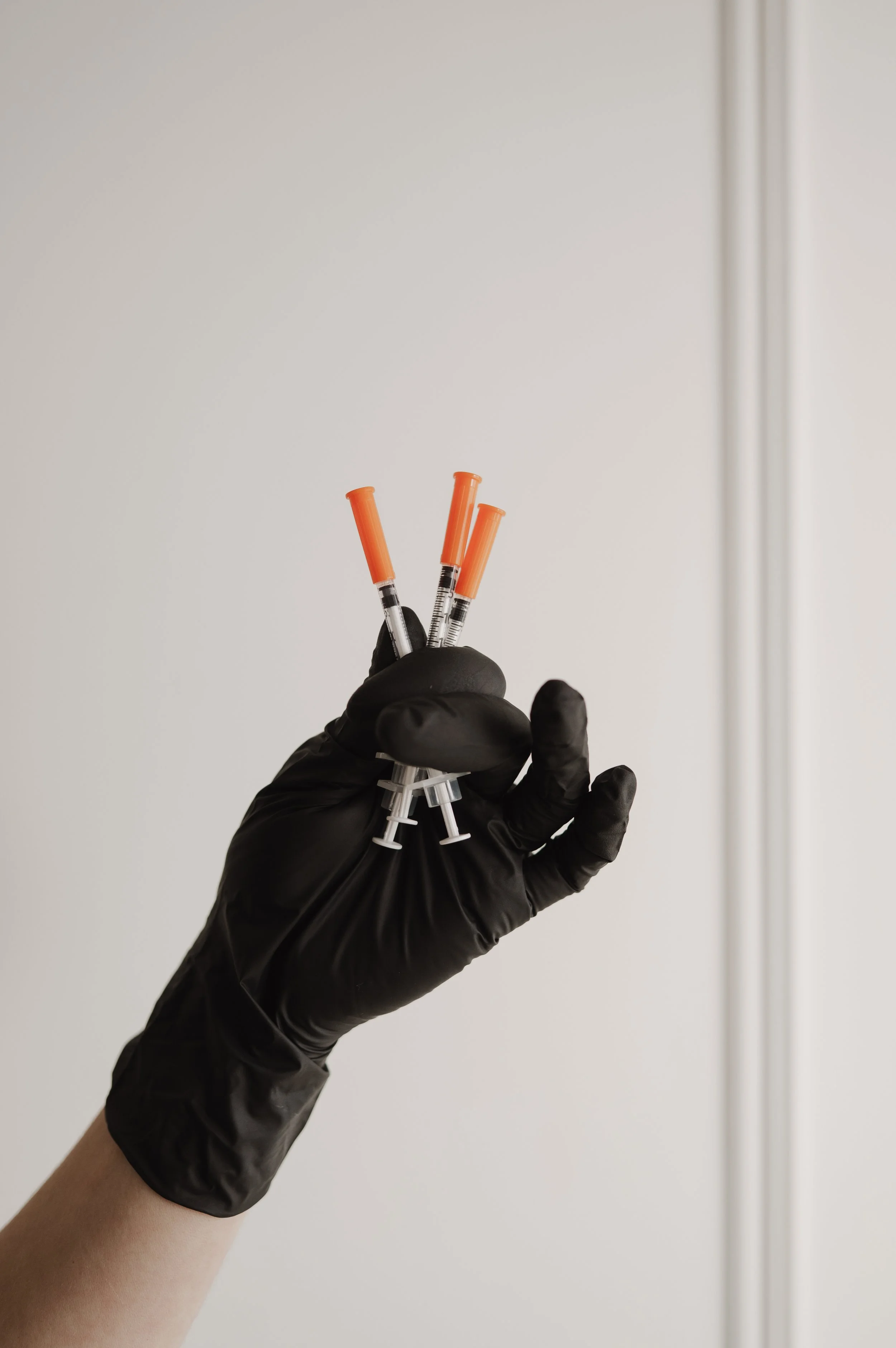 A gloved hand holding three syringes filled with a clear liquid in front of a plain white wall.