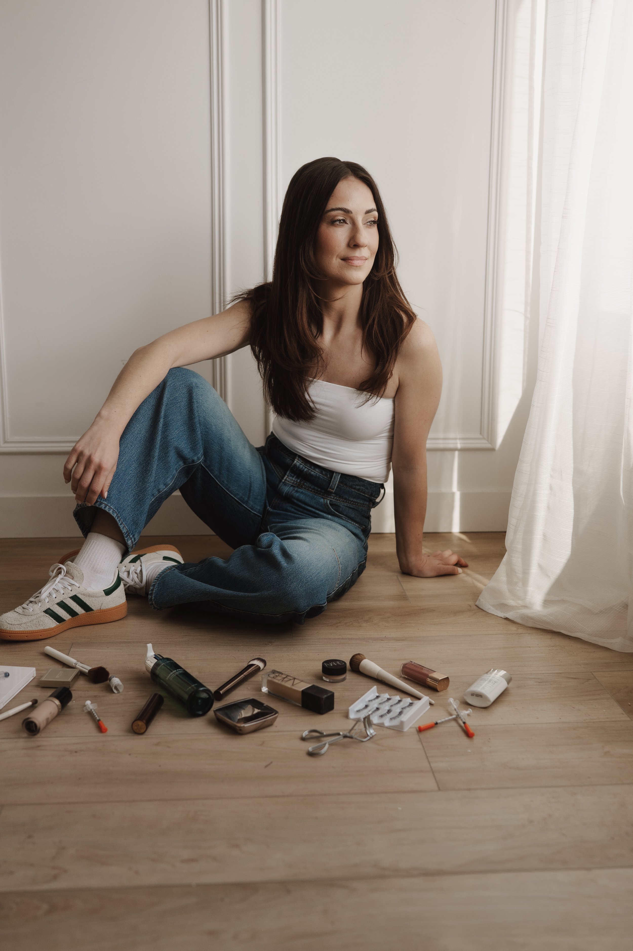 A woman with long brown hair, sitting on the floor next to a white wall with wainscoting, surrounded by makeup and skincare products, looking out the window with sheer white curtains.