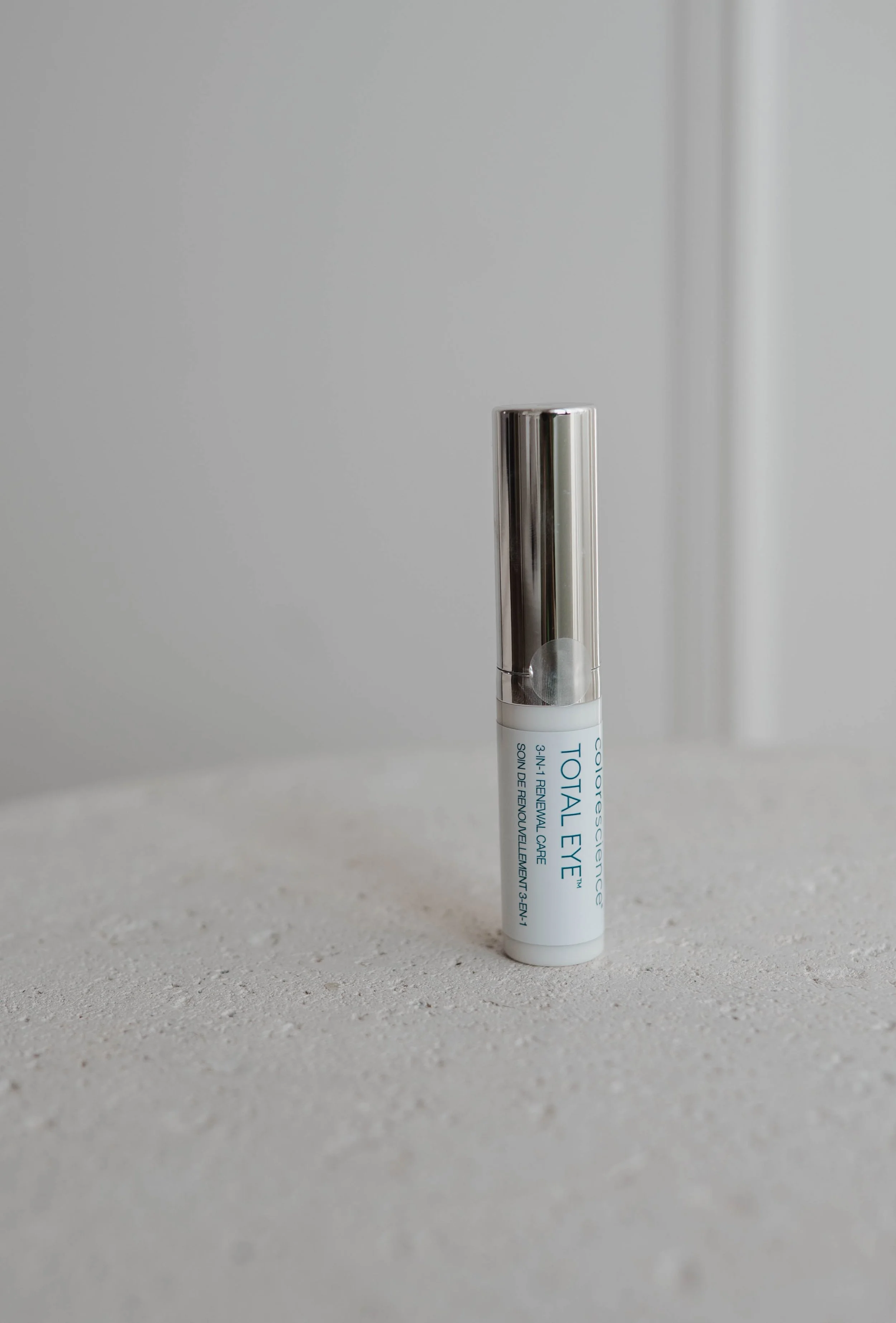 A white cylindrical container of Total Eye Renewal Care with silver cap standing on a light gray textured surface, against a plain light gray background.