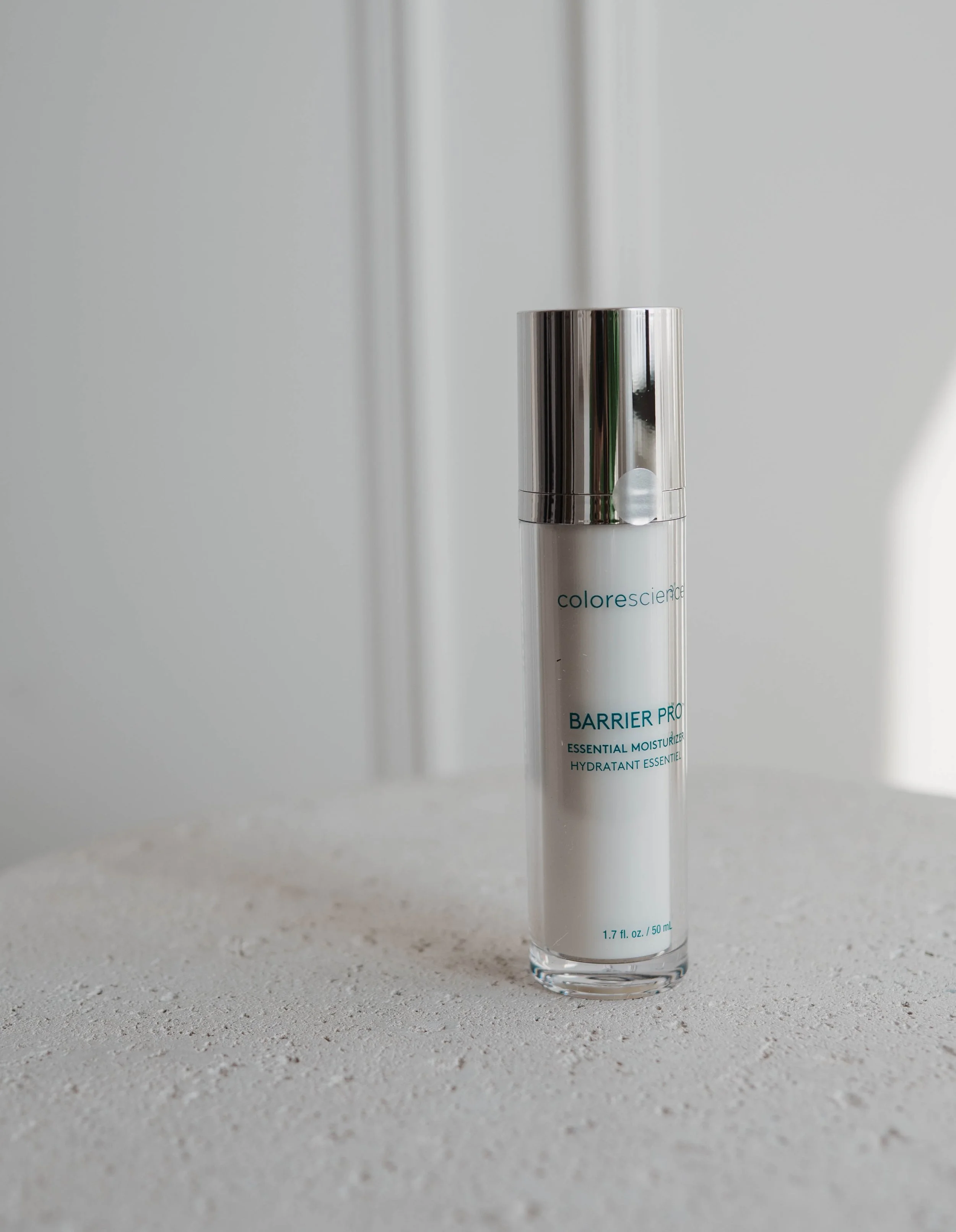 A white cylindrical bottle of skincare product with a silver cap, labeled 'Barrier Pro Essential Moisturizer,' placed on a light textured surface with a plain background.