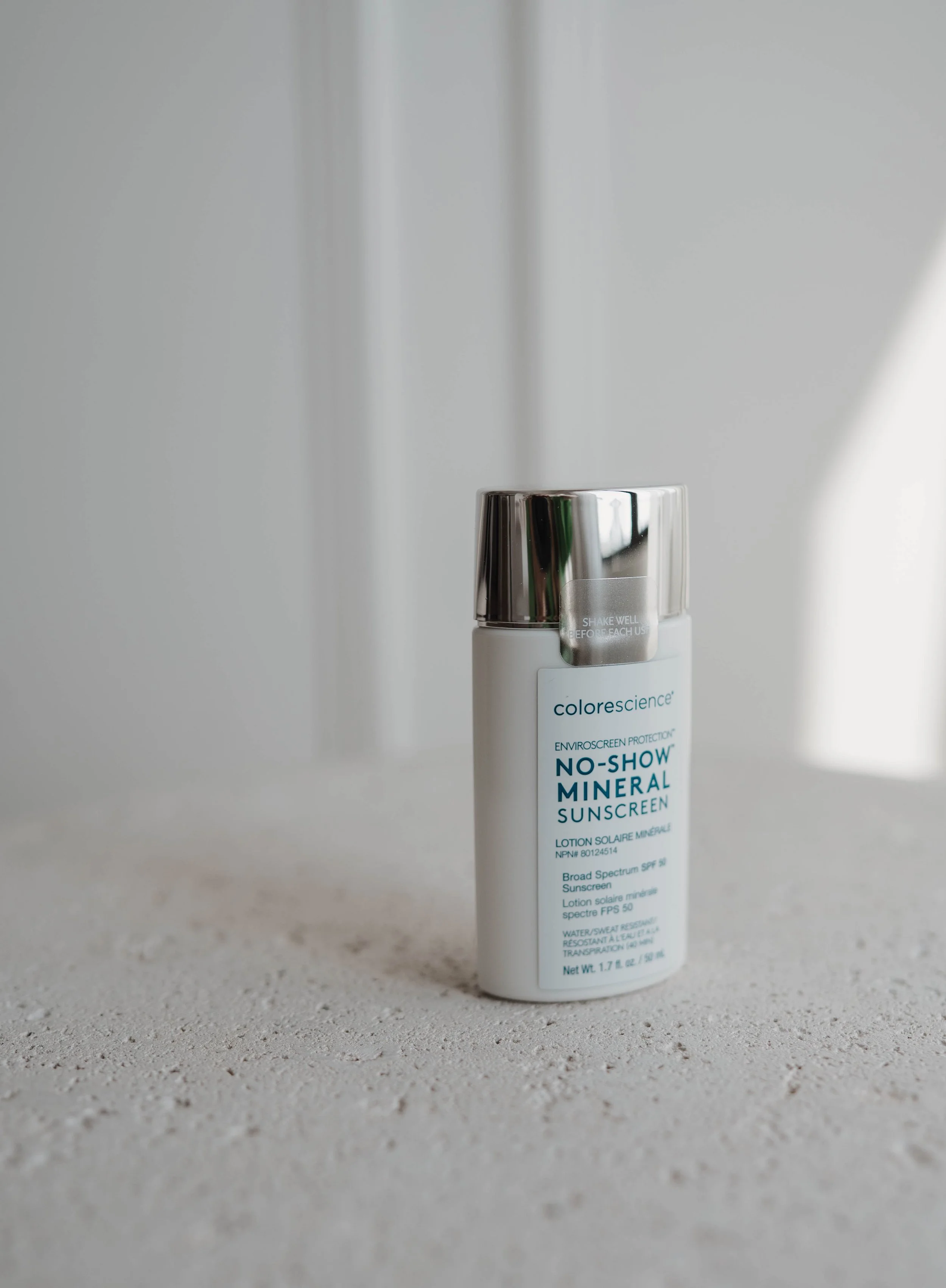 A white bottle of Colorescience No-Show Mineral Sunscreen with a metallic cap, placed on a textured surface against a blurred gray background.