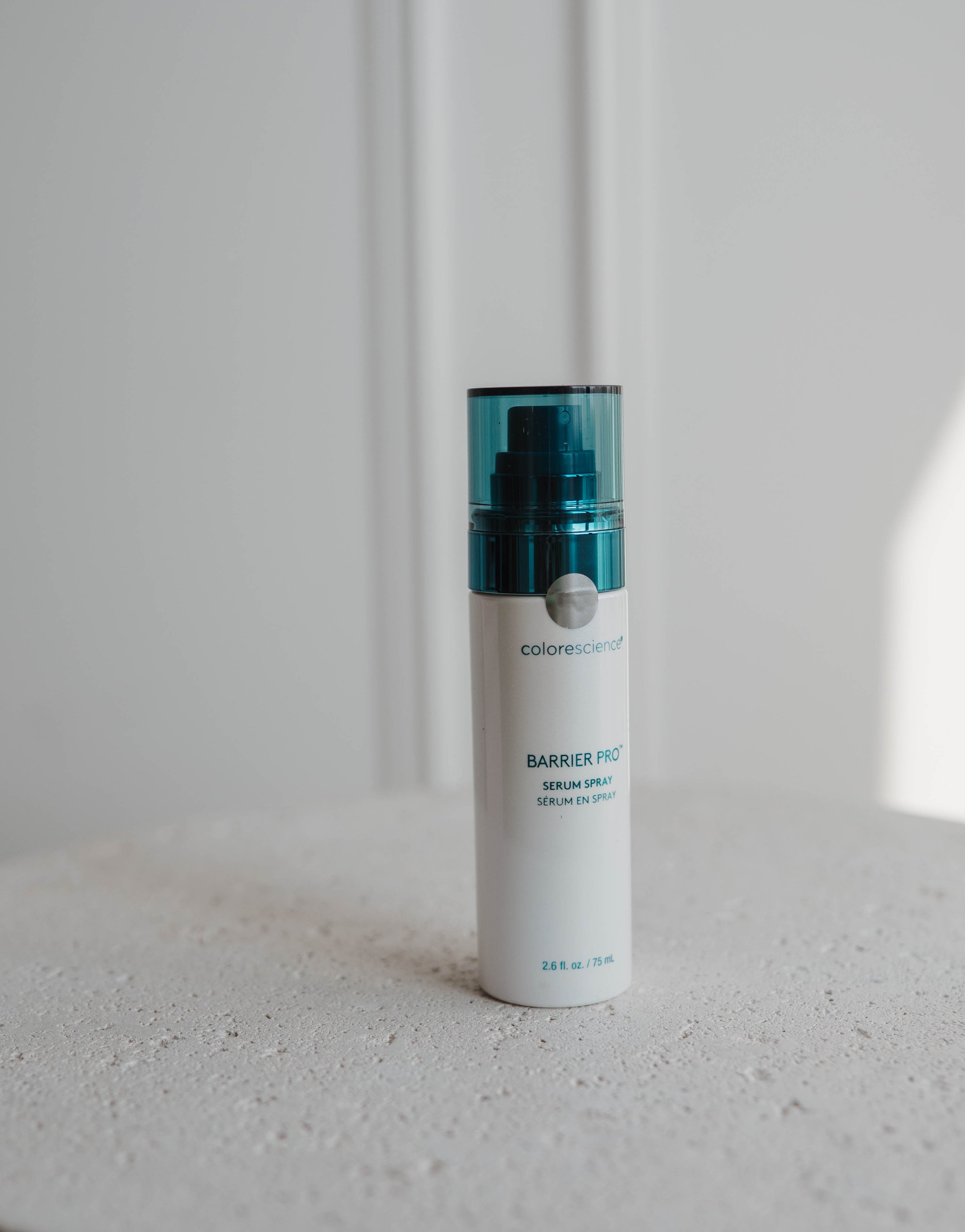 A white bottle with a blue pump labeled 'Barrier Pro Serum Spray' standing on a textured surface with a plain background.