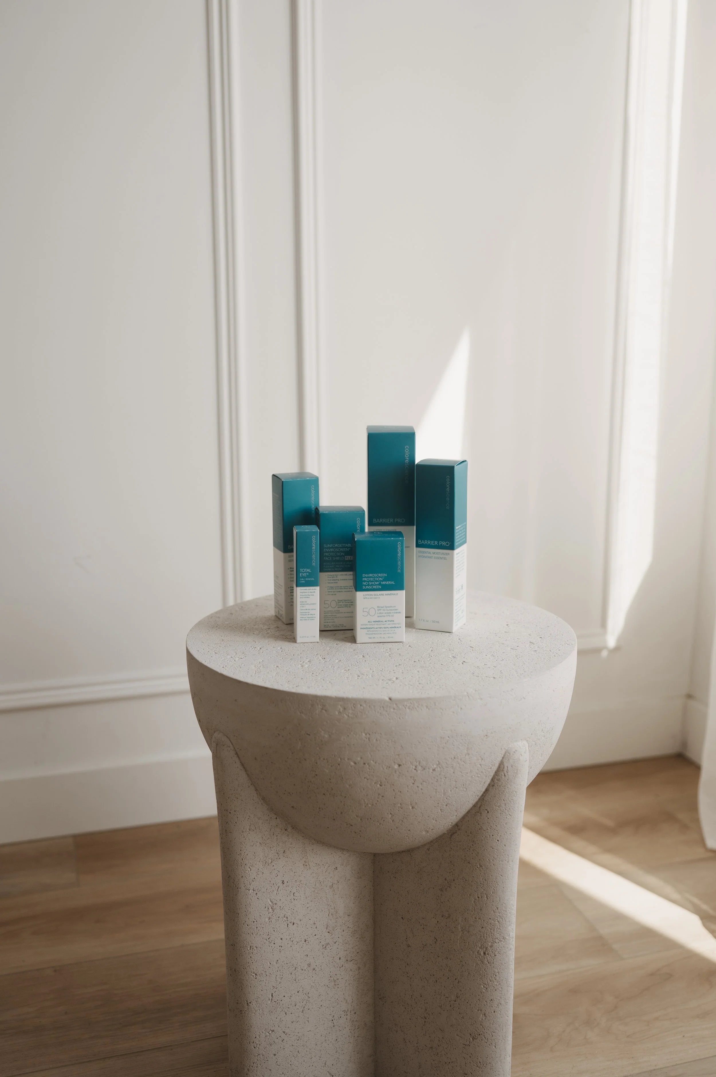 A white, textured, stone-like round table with a short, thick pedestal base. On top, there are several blue and white skincare product boxes grouped together. The background includes white paneled walls and light wooden flooring, with natural sunlight casting shadows.
