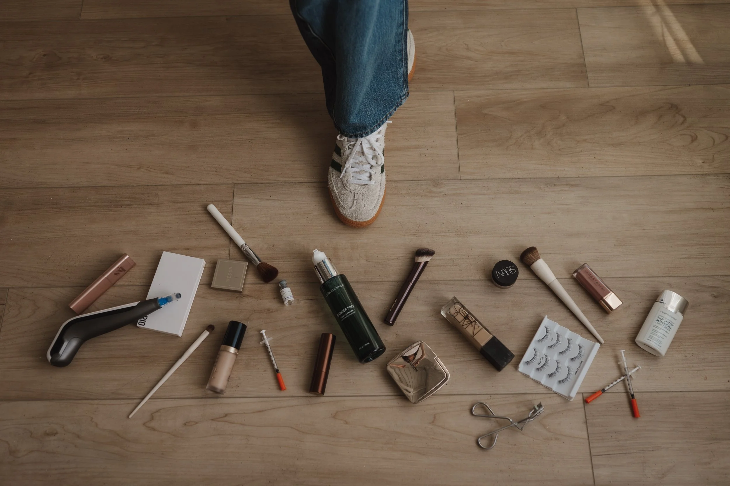 A person's leg wearing blue jeans and a white sneaker with green stripes, standing on a wooden floor surrounded by various makeup and beauty products such as brushes, lip gloss, mascara, and skincare bottles.