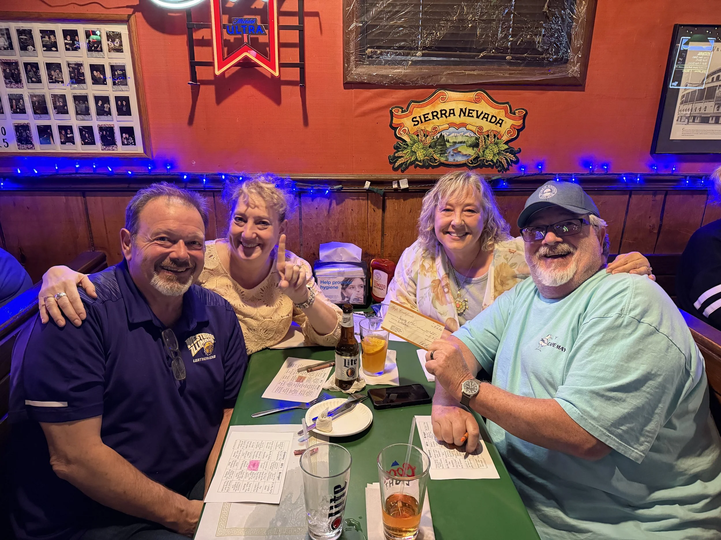 Four people sitting at a table in a restaurant, smiling and posing at the camera. The table has drinks, menus, and utensils, with a red wall, blue string lights, and framed photos and signs on the wall in the background.