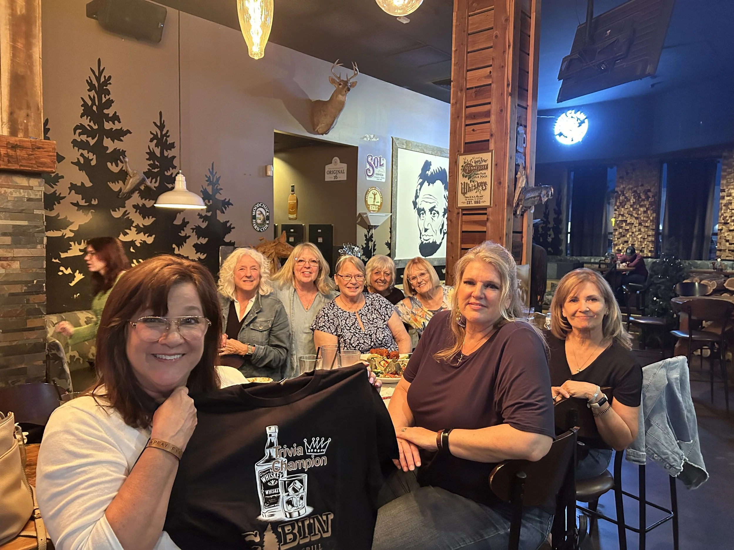 A group of women sitting at a restaurant table, smiling and posing for the photo. The restaurant has rustic decor with wall murals of trees, a mounted deer head, and framed pictures. One woman in the foreground wears glasses and a light-colored shirt, and has a black bag with white text. The group is gathered for a meal, and the atmosphere appears casual and friendly.