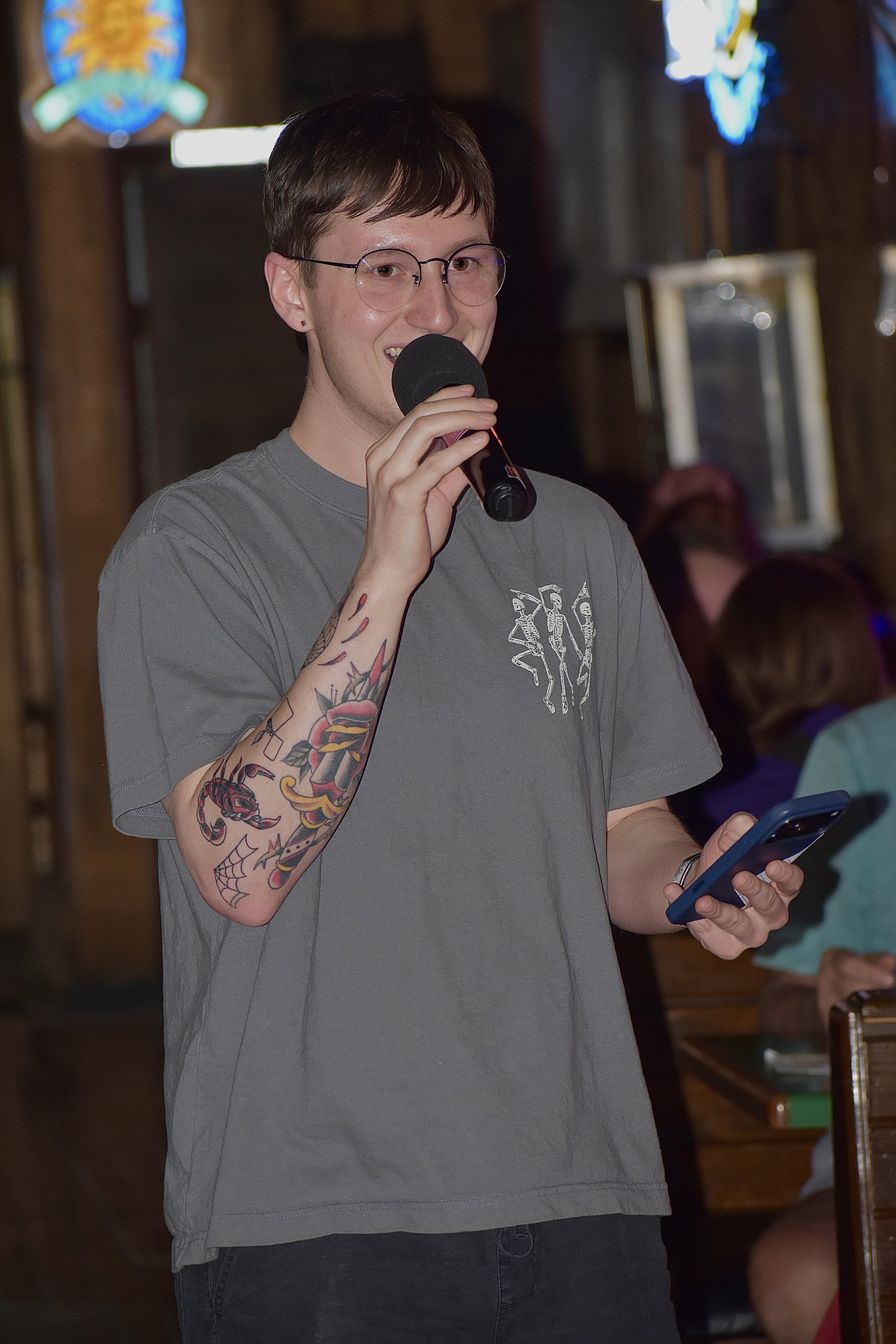 A young man with glasses and short brown hair, holding a microphone in his right hand and a smartphone in his left, standing indoors with stained glass windows in the background.