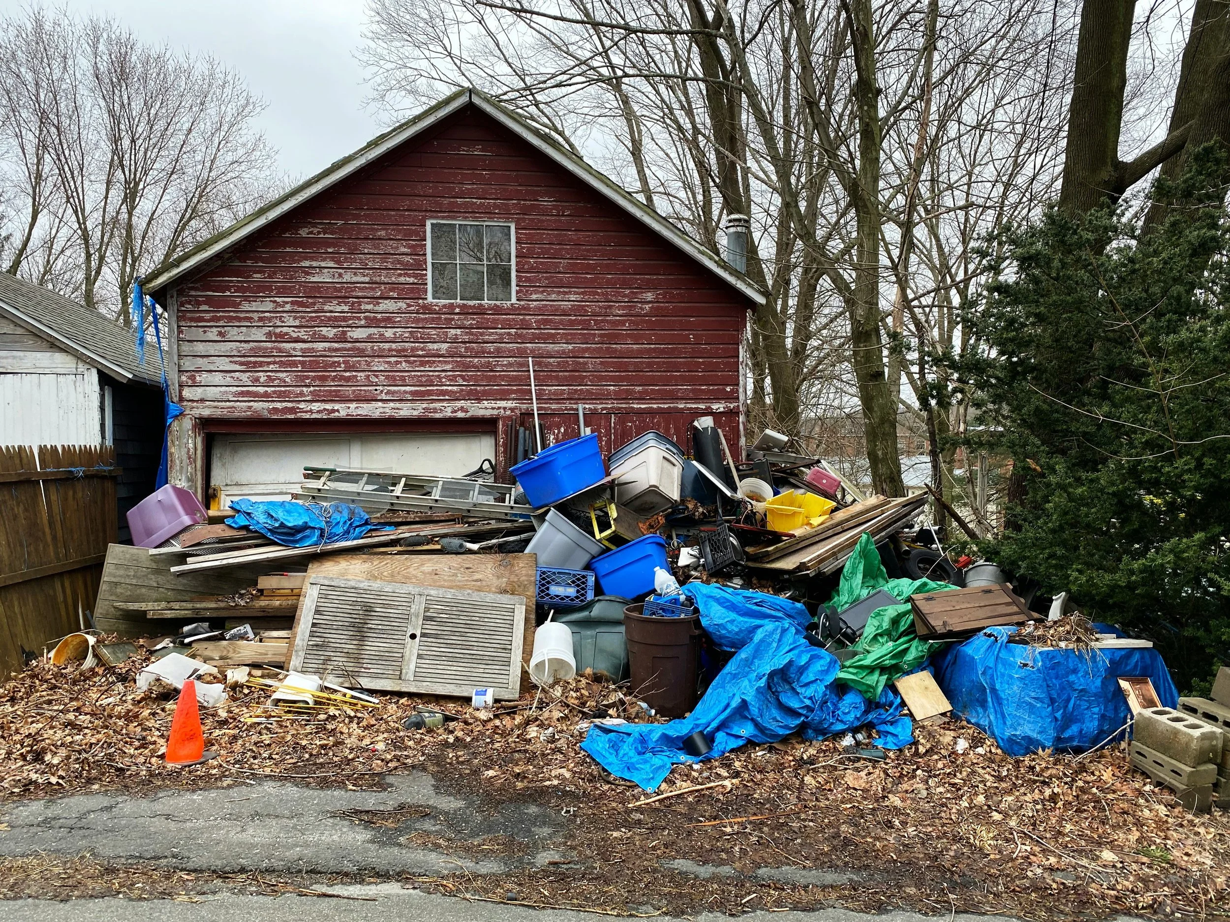 Photo of an old red home with paint peeling off and a broken white garage door. Junk is overflowing in front of it.