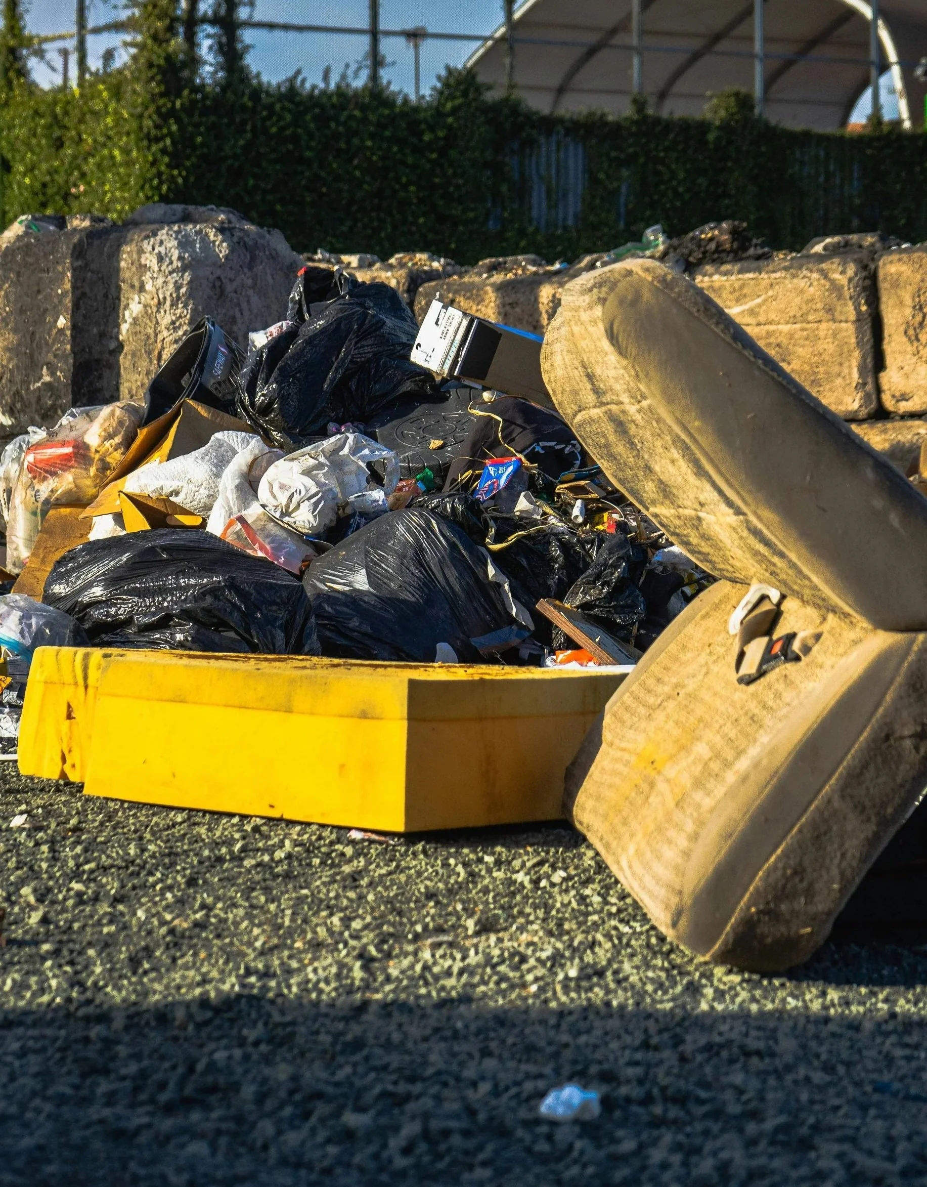 Pile of trash including black plastic bags, cardboard boxes, and miscellaneous litter, against a stone wall and a partly visible structure in the background.