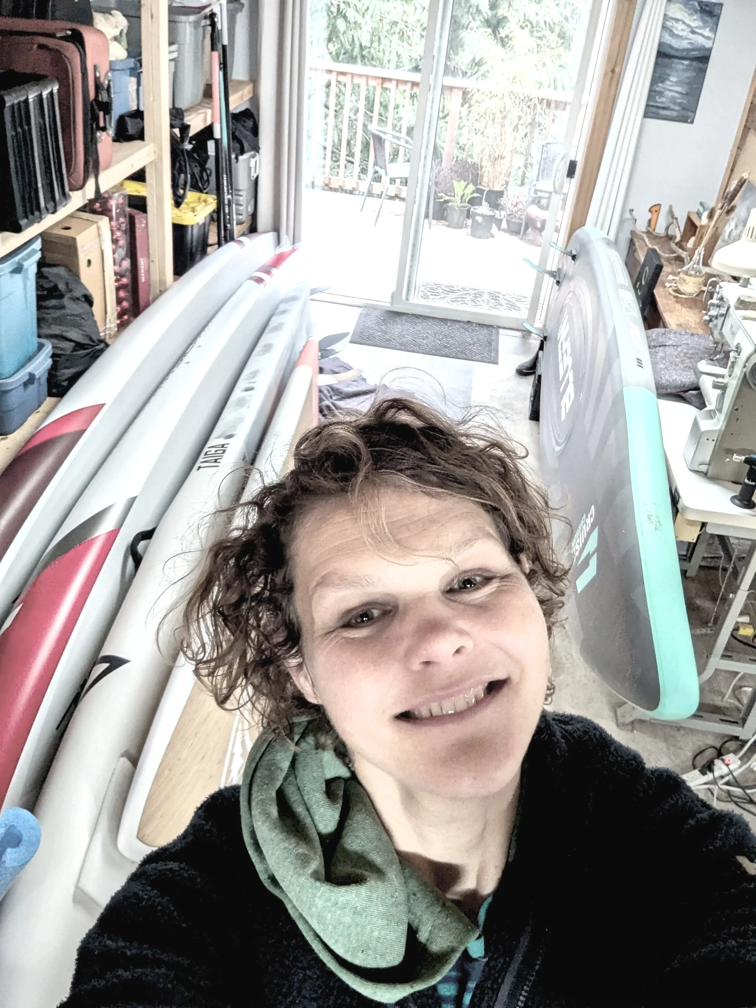 A person taking a selfie in a room with surfboards and a view of a patio outside.