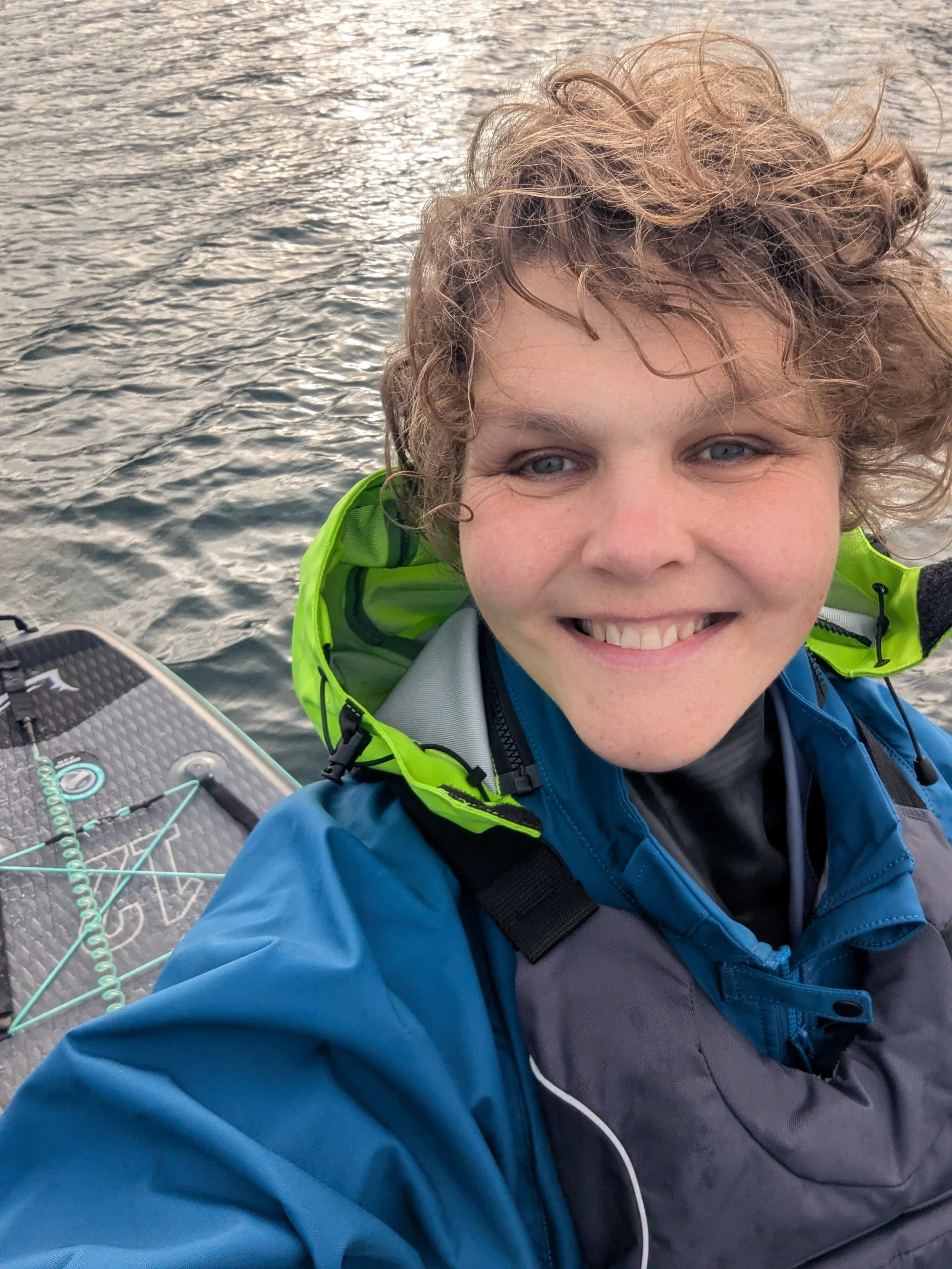 A person taking a selfie near water, wearing a blue jacket with a bright green hood, smiling, with part of a paddleboard visible.