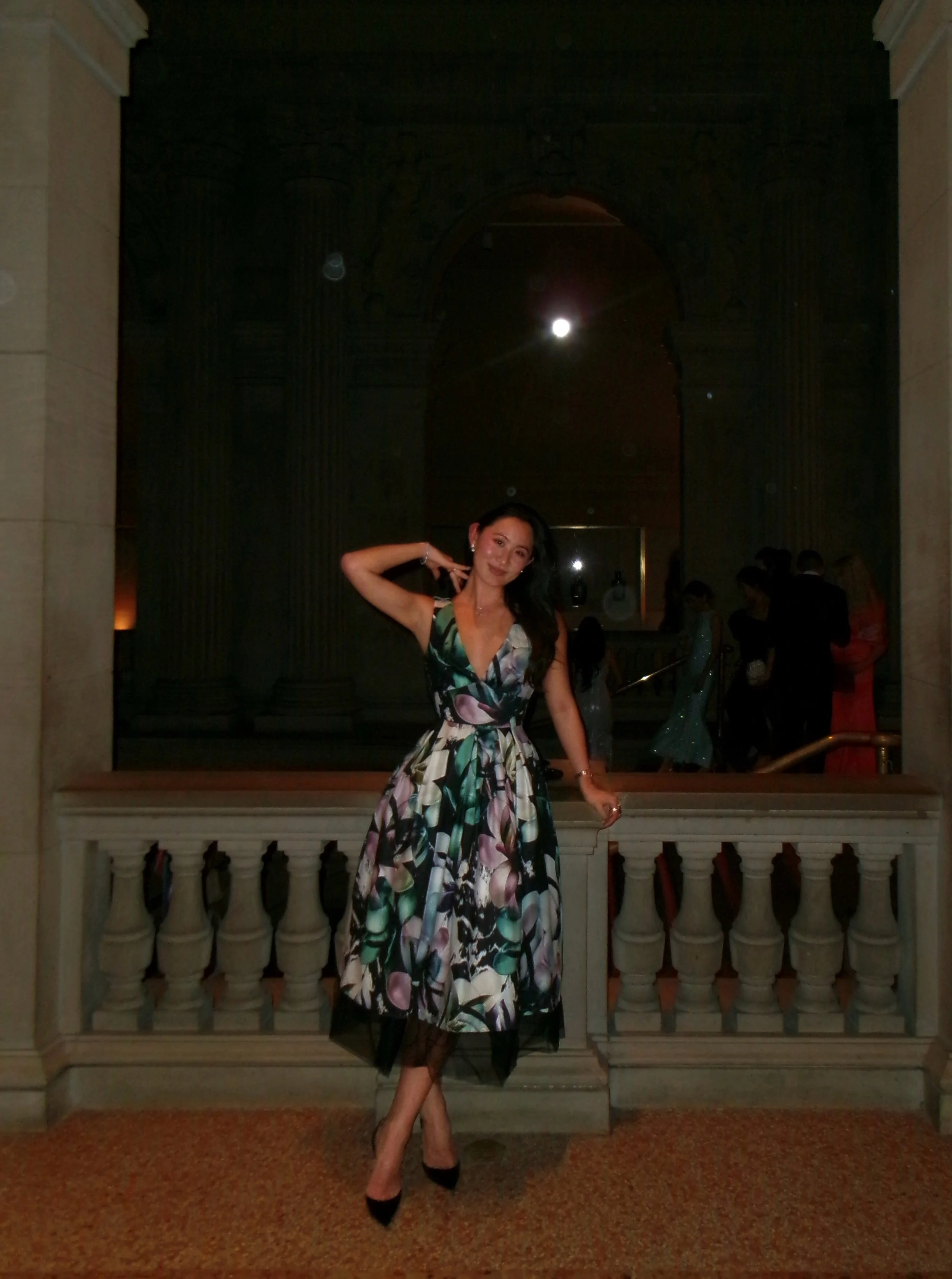 A woman in a floral dress posing on a balcony with a marble balustrade, in an elegant indoor setting with ornate architectural details and other people in the background.
