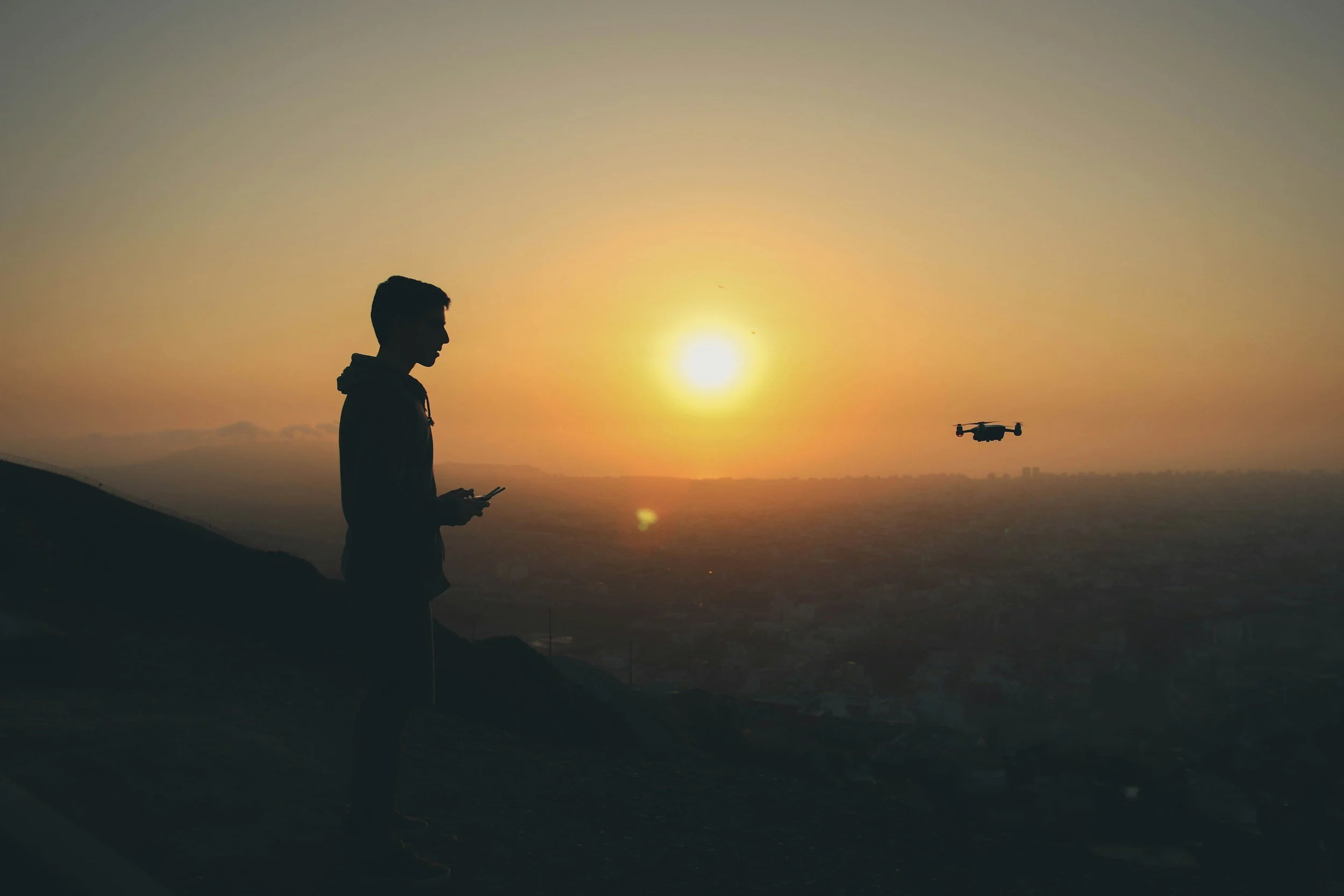 Silhouette of a person looking at a drone flying during sunset over a landscape.