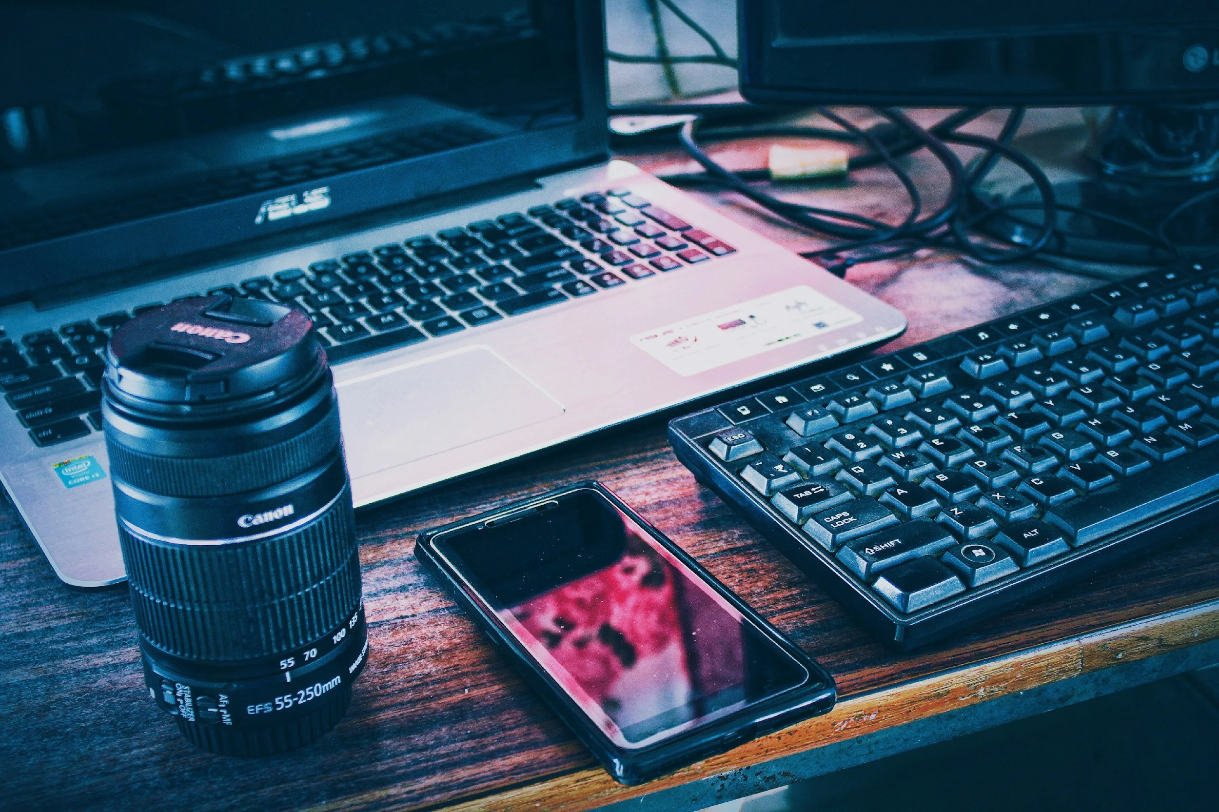 A cluttered wooden desk with electronic devices including a laptop, external keyboard, smartphone, and a Canon camera lens. There is a small container and tangled cables on the desk.