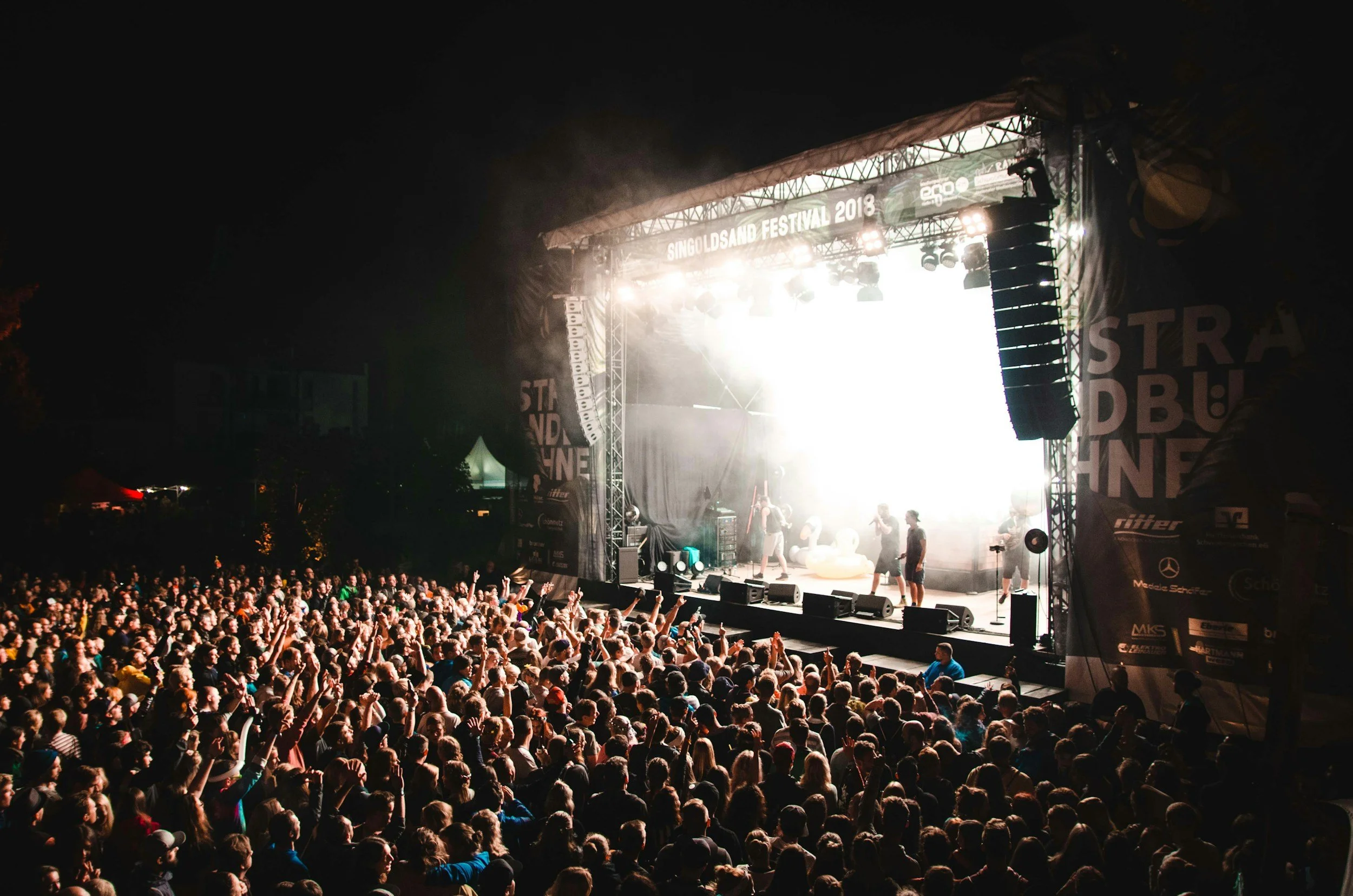 A large crowd attending a concert at night with a brightly lit stage displaying banners for the Singoldsand Festival 2018. People in the audience are facing the stage, raising their hands, and enjoying the performance.