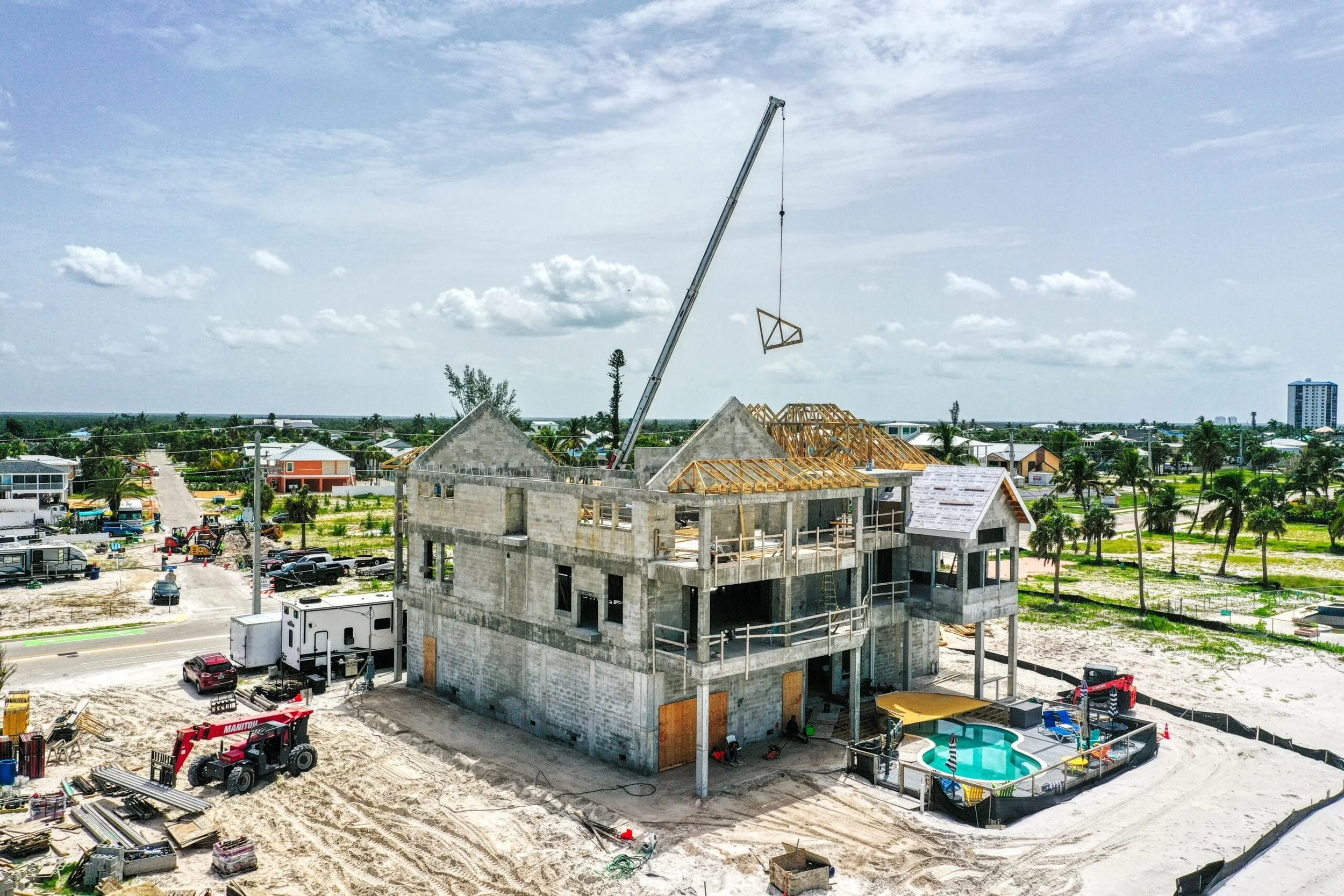 Construction site with partially built concrete house, construction crane lifting a wooden roof truss, and surrounding equipment, vehicles, and neighboring houses under a partly cloudy sky.