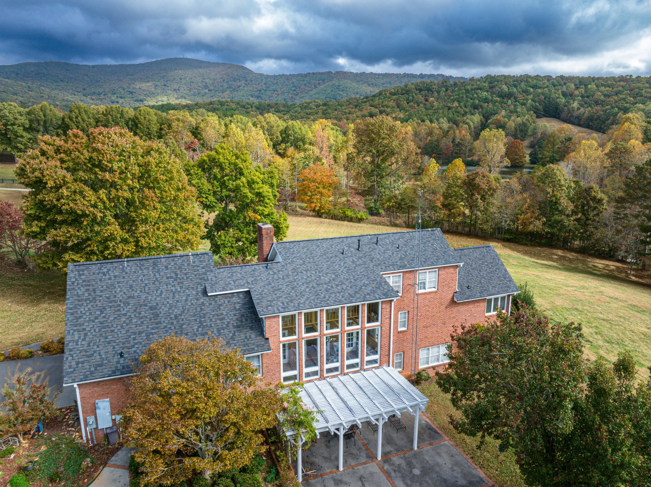 An aerial view of a brick modern house with large windows, surrounded by green trees with some leaves turning colors and a gray tiled roof. In the background, there are rolling forested hills under a dark, cloudy sky.