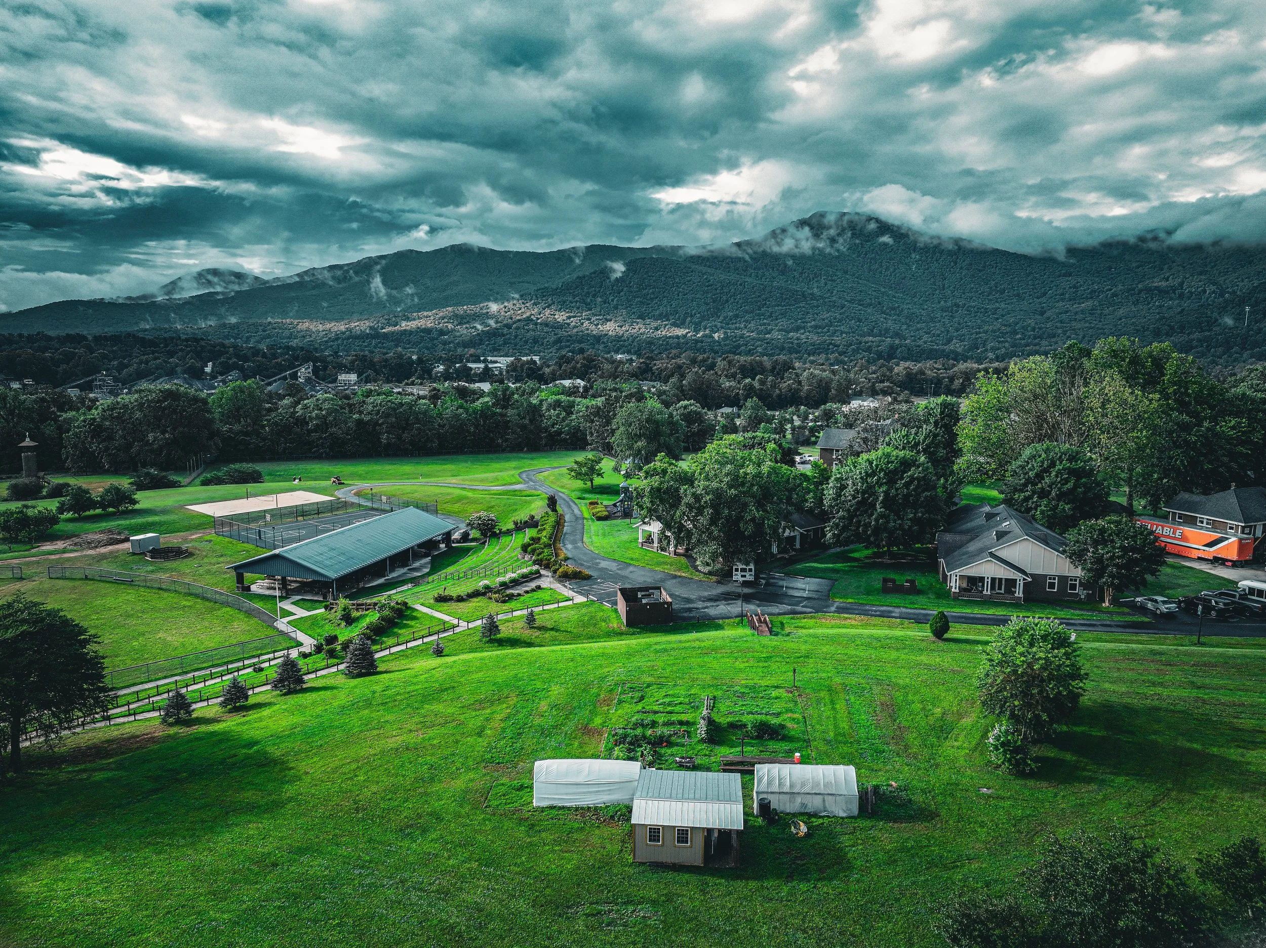 A scenic view of a mountainous landscape with a park, sports courts, trees, and residential houses in the foreground. The sky is cloudy with patches of mist around the mountain peaks.