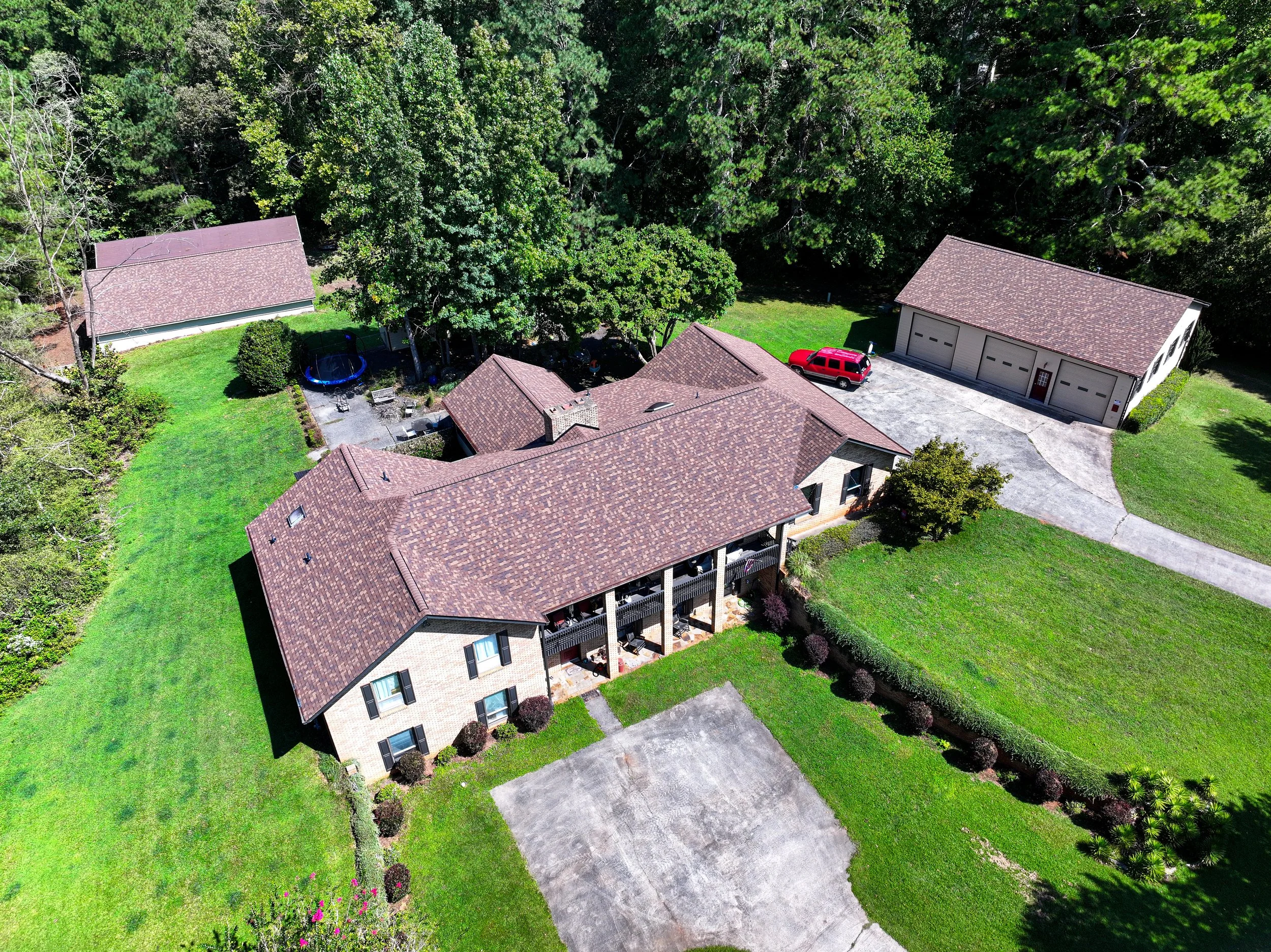 Aerial view of a residential backyard with a brick house, a trampoline, a red SUV, a garage, and surrounded by lush green trees and grass.