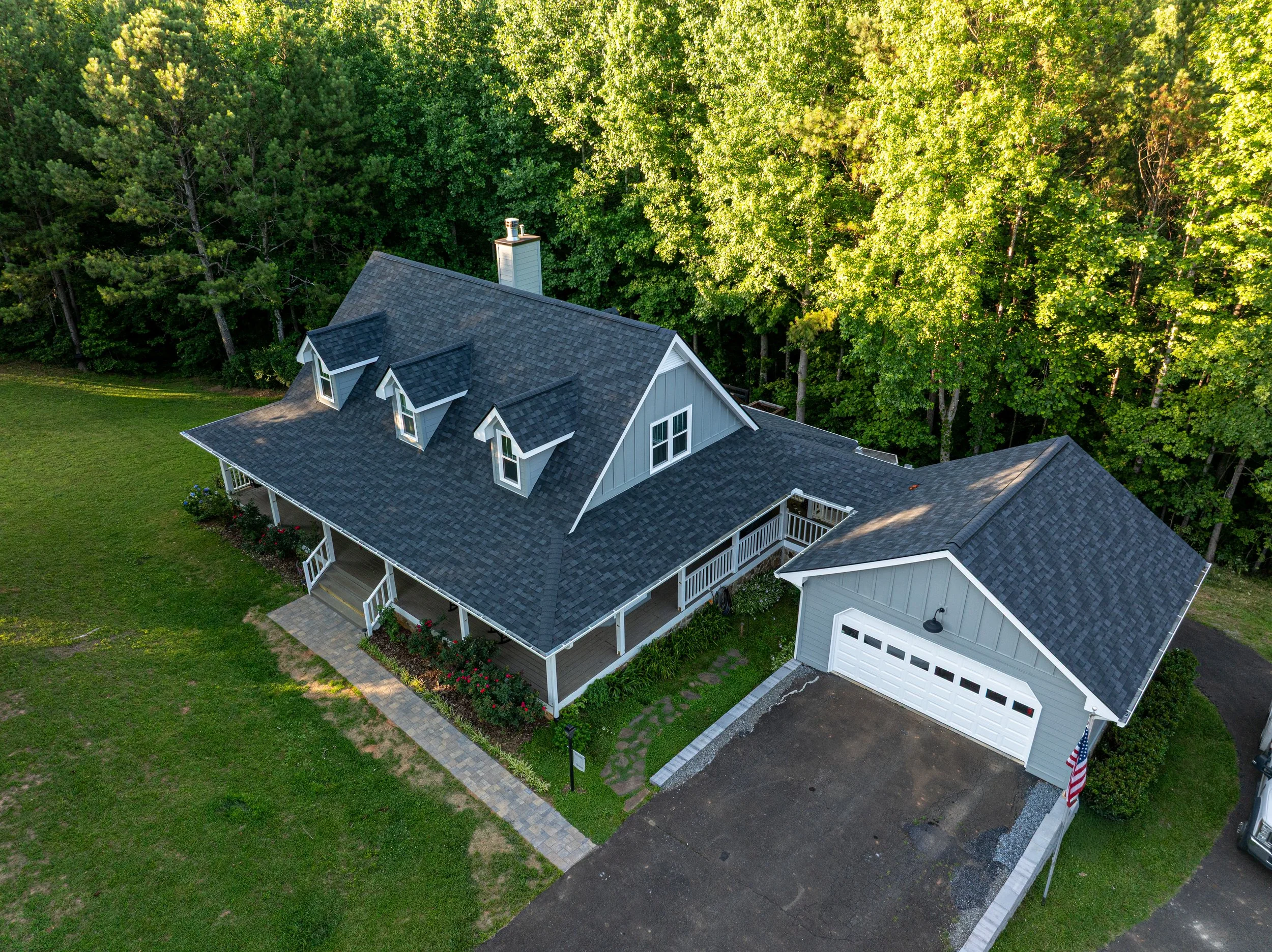 An aerial view of a house with gray roofing, white siding, a front porch, and a adjacent garage. The house is surrounded by lush green trees and a well-maintained lawn.