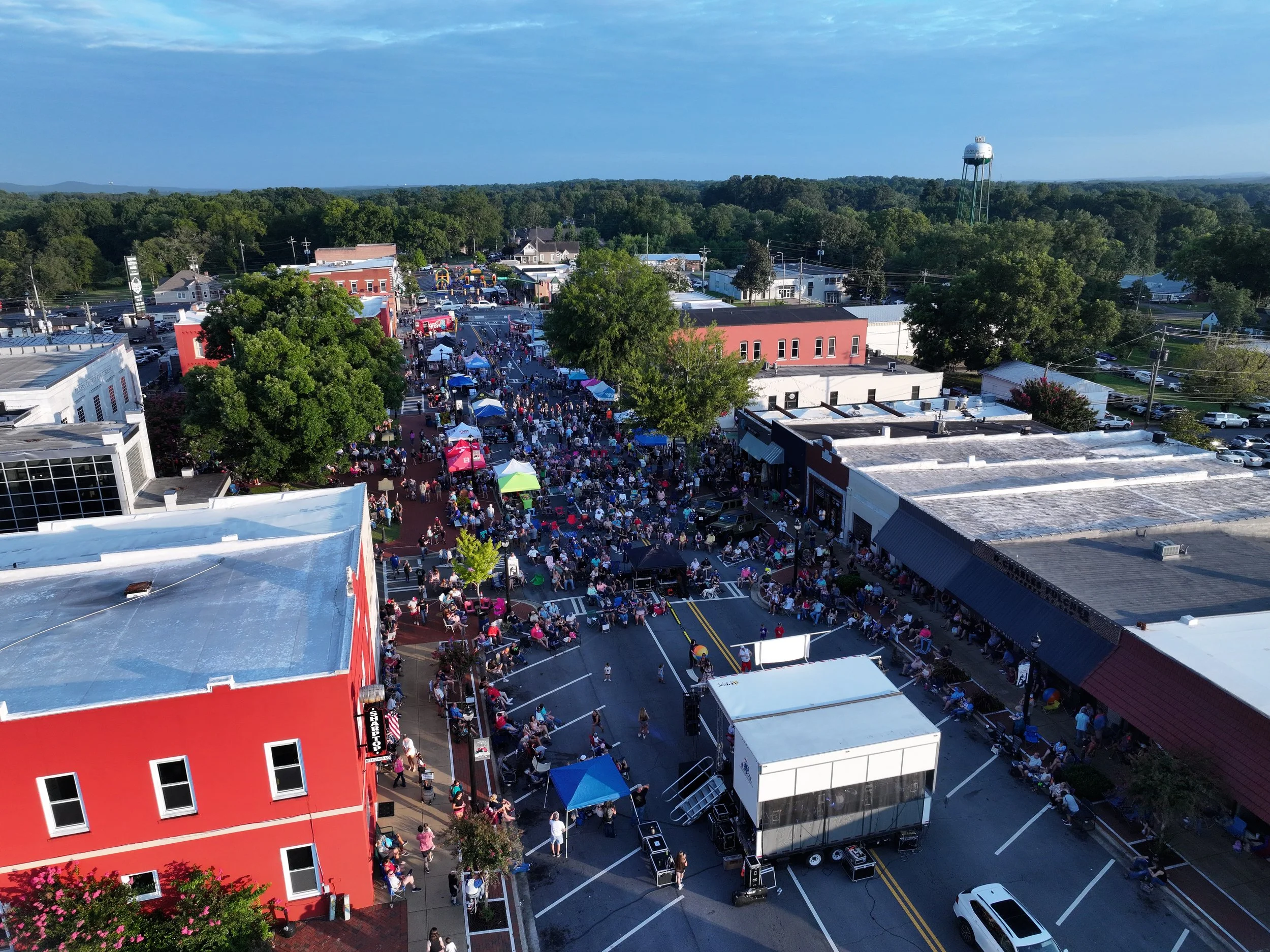An aerial view of a lively outdoor event in a small town, with numerous tents, booths, and a large crowd of people gathered in a downtown area surrounded by trees and buildings, and a water tower in the distance.