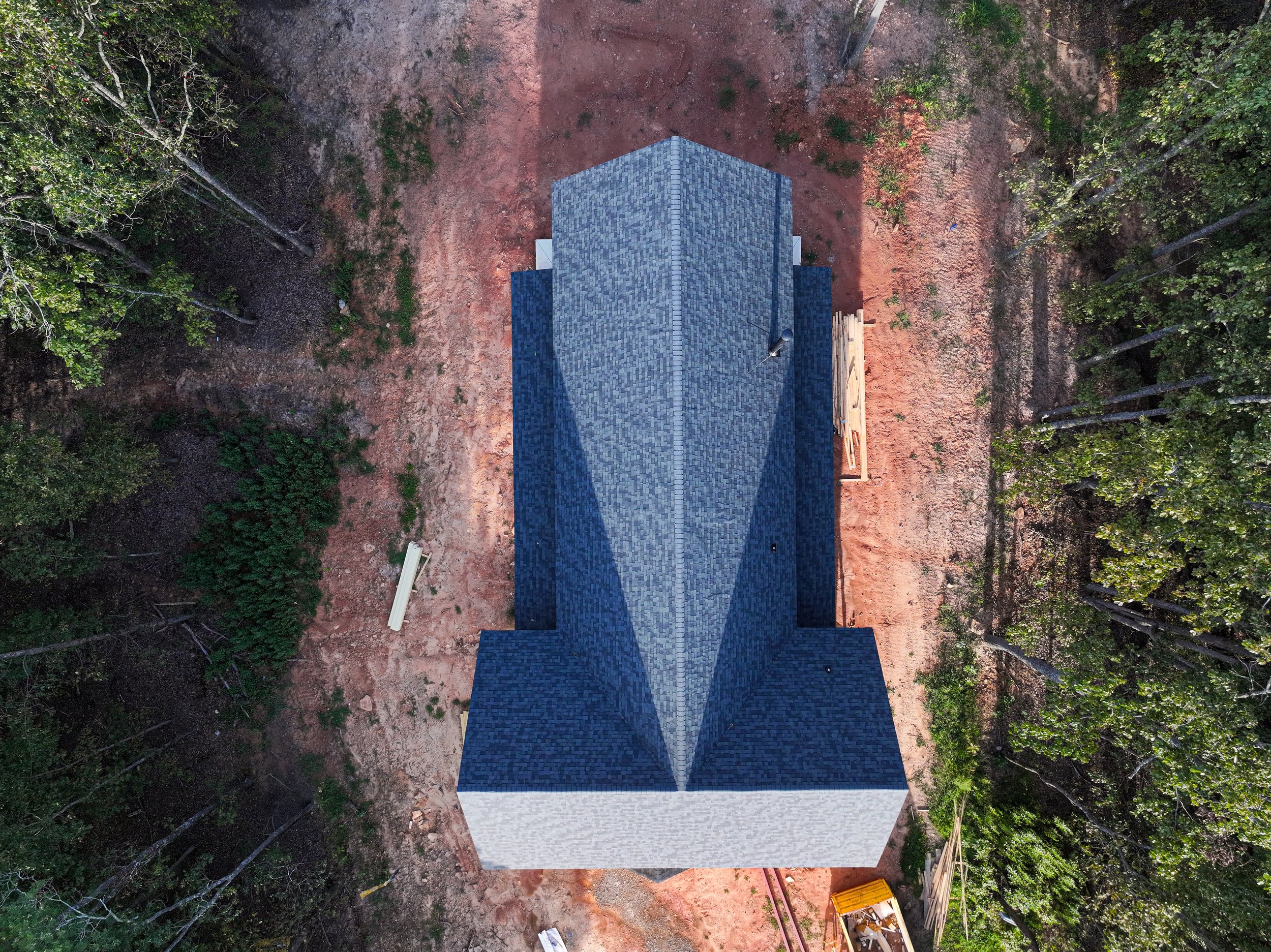 Aerial view of a house with a dark gray shingle roof, surrounded by trees and cleared land.