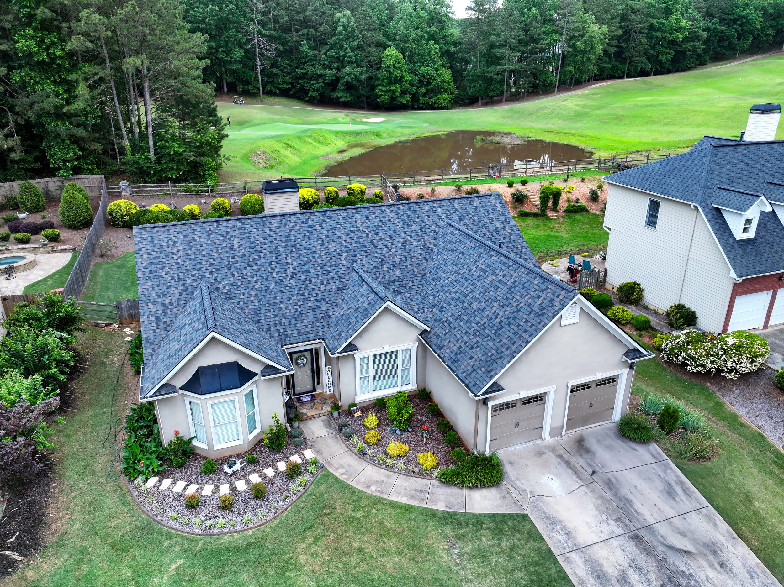 A suburban house with a dark gray shingle roof, white exterior walls, and a well-maintained front yard with bushes and flowers. There is a paved driveway leading to a garage, and the front door is visible. The backyard has a fenced area, a small gard