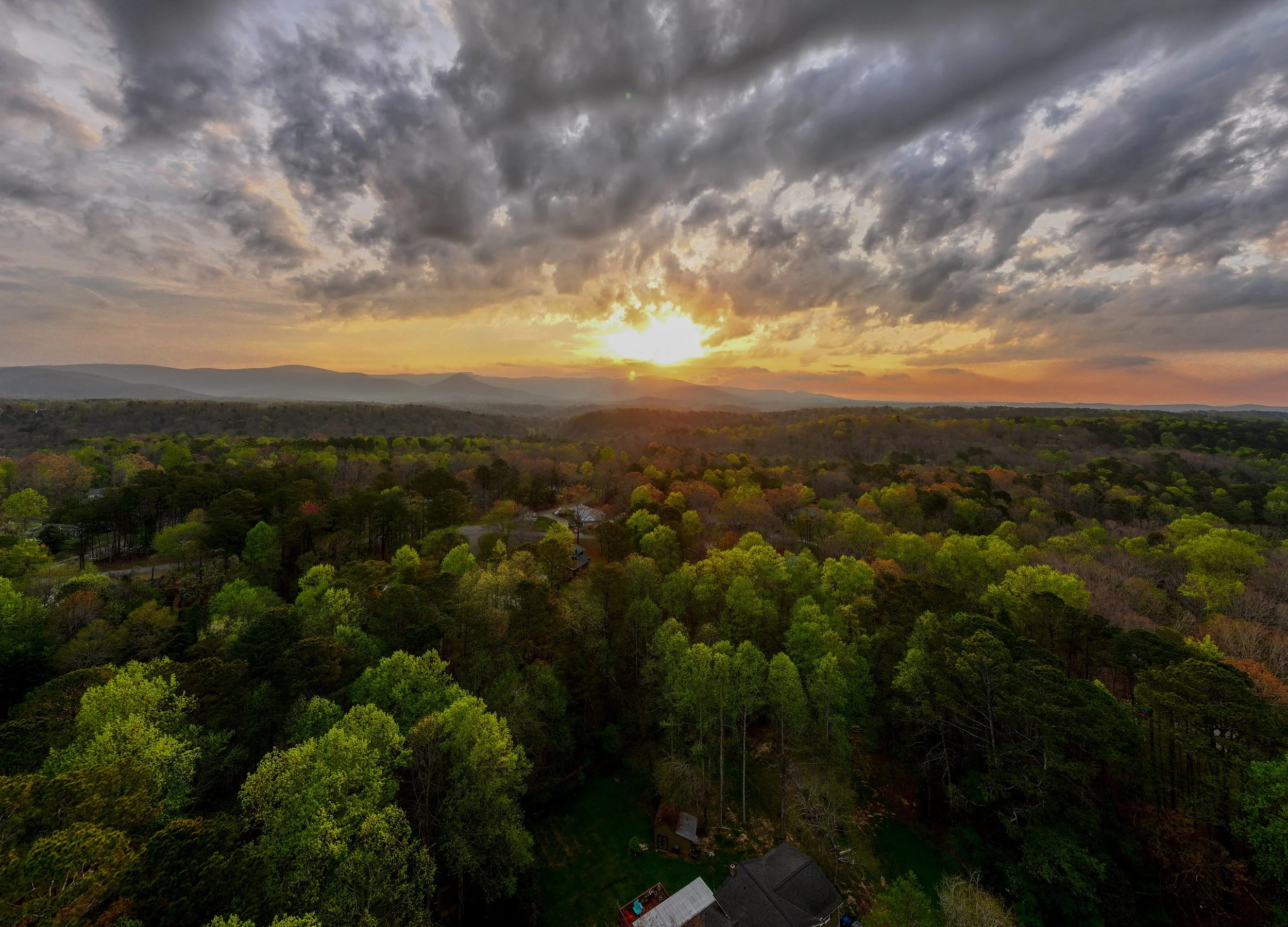 A scenic view of a sunset over a valley with a cloudy sky, distant hills, and a dense forest in the foreground.
