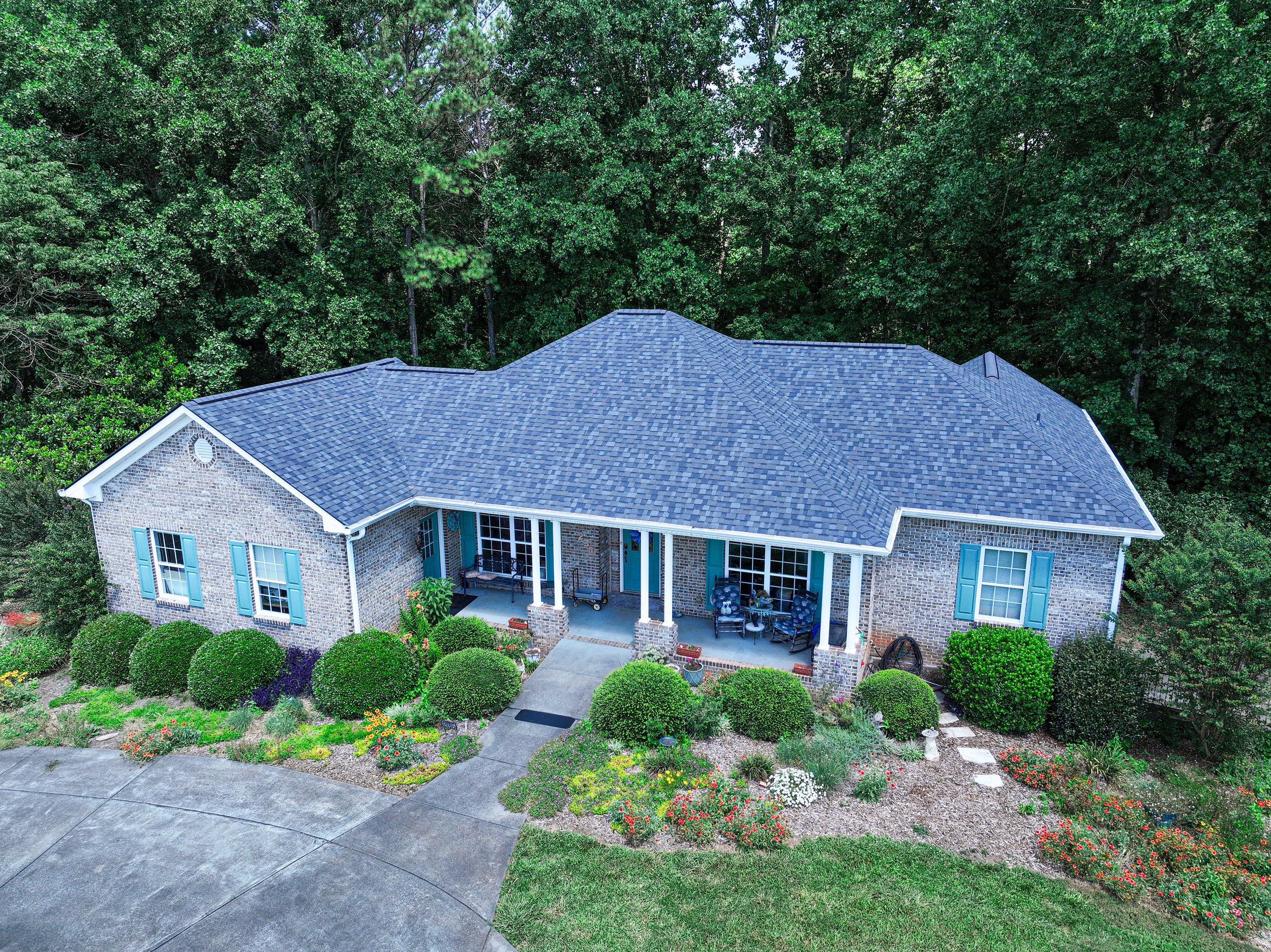 A house with a gray-shingled roof surrounded by lush green trees and well-maintained flower beds.