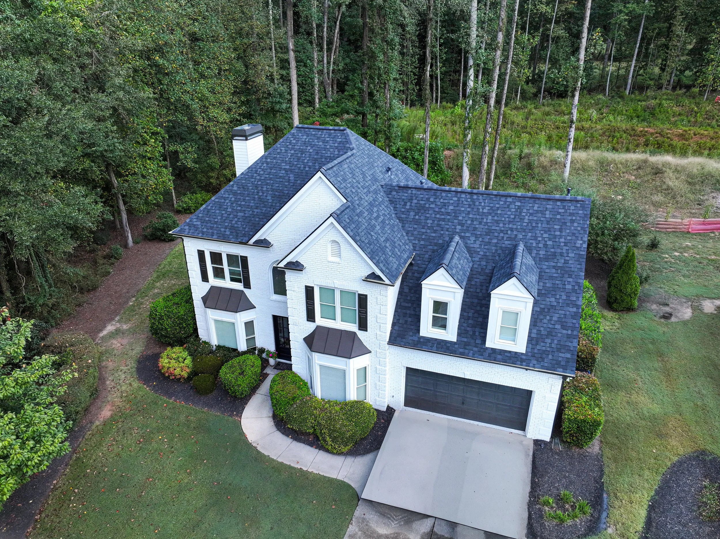 A white two-story house with black trim and a dark shingled roof, surrounded by trees and a well-maintained lawn with bushes and a curved sidewalk.