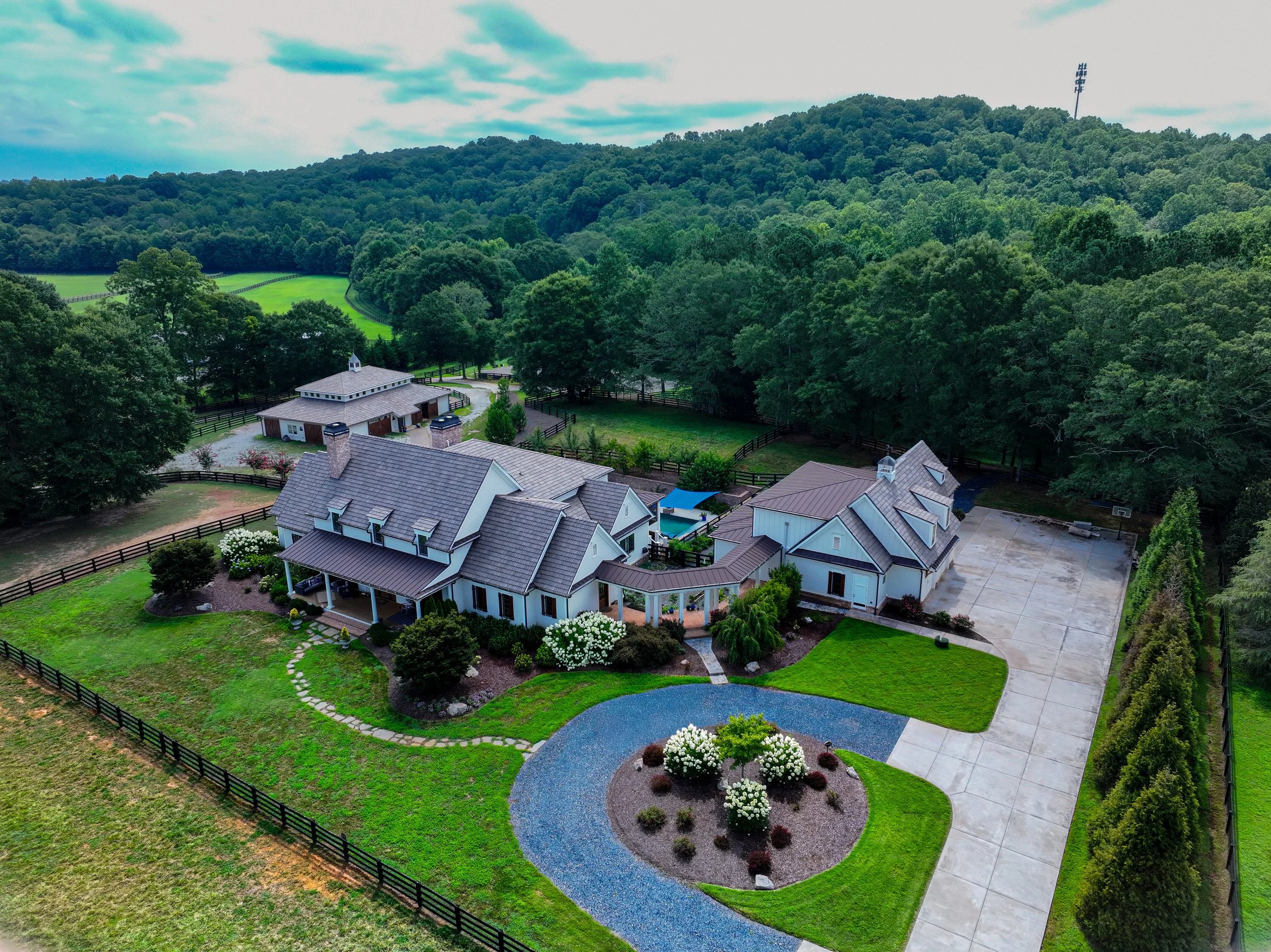 Aerial view of a residential neighborhood with houses, landscaping, and a backyard pond, surrounded by green trees and forested hills under a partly cloudy sky.