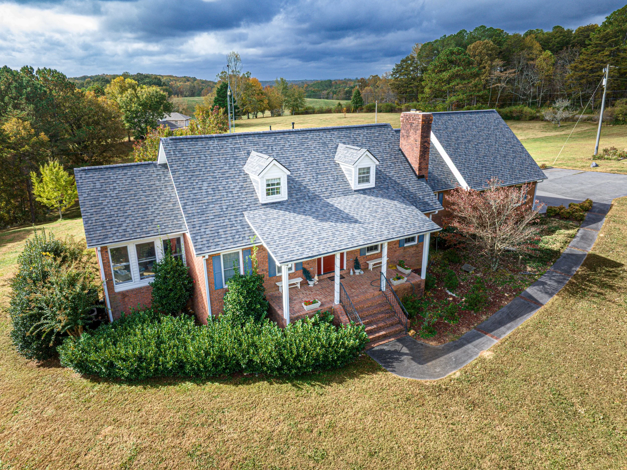 An aerial view of a brick house with a gray shingle roof, surrounded by trees and a lawn. The house has dormer windows, a brick chimney, and a brick patio with steps leading to the front door. There is a parking area and a paved walkway near the house, with a backdrop of open fields, trees, and a cloudy sky.