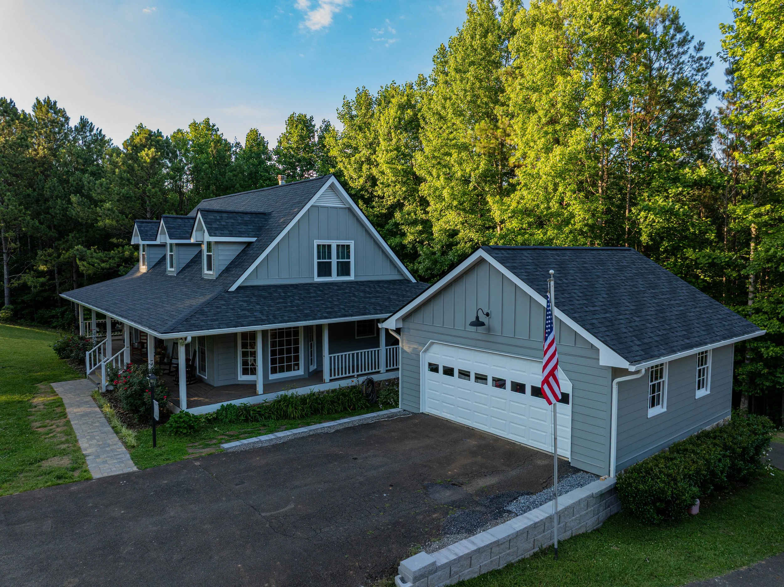 A two-story house with grey siding, white trim, and a gabled roof situated among lush green trees under a clear blue sky.