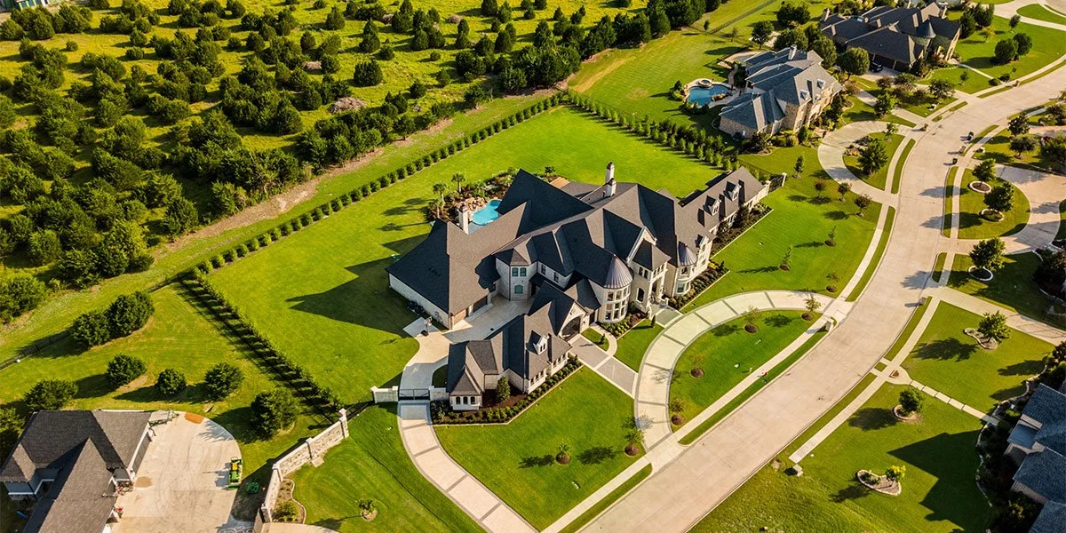 An aerial view of a large, luxurious house with a spacious yard, surrounded by well-manicured lawns, trees, and neighboring houses in a suburban neighborhood.
