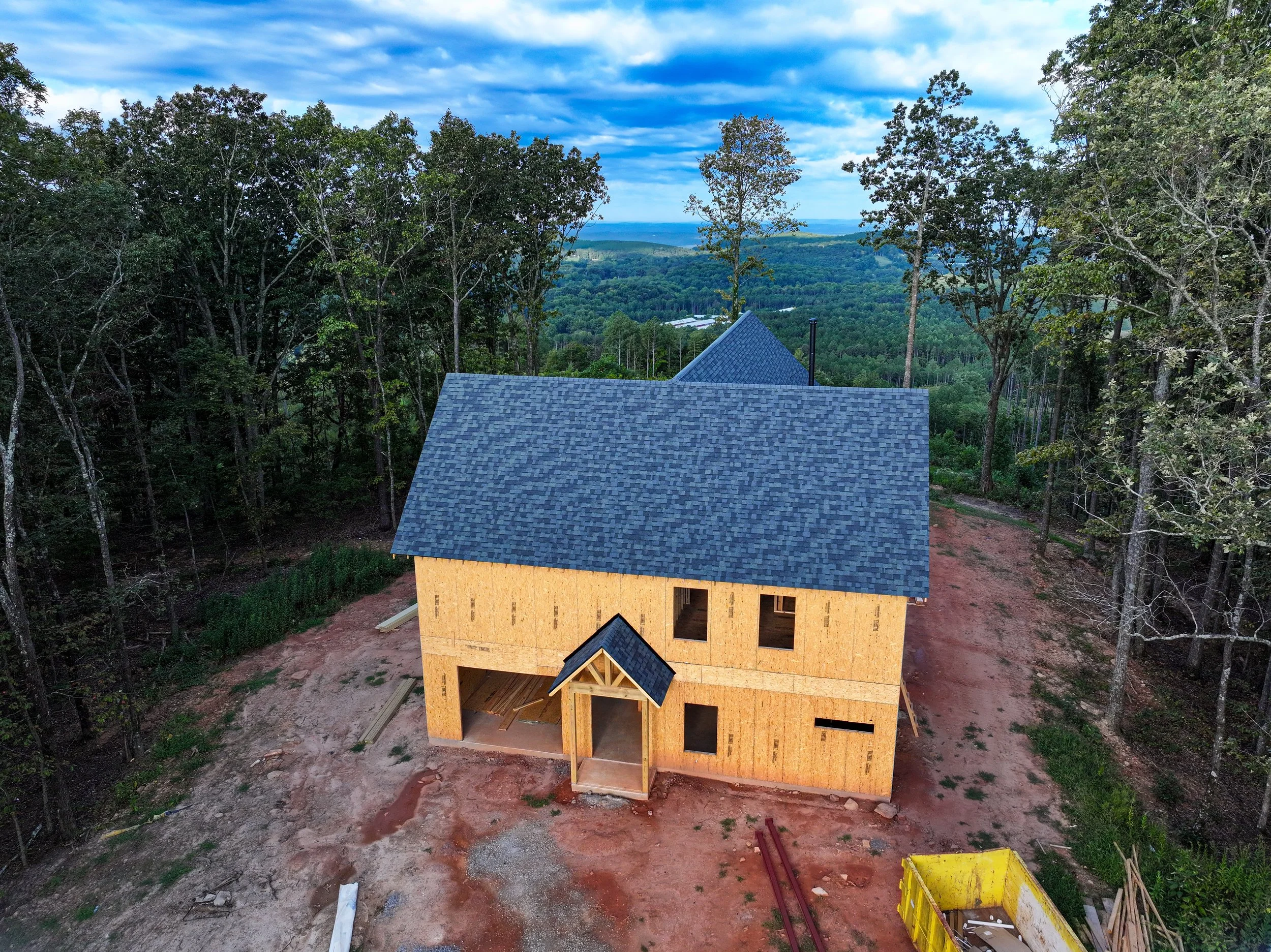 A house under construction in a wooded area with a view of hills and a blue sky with scattered clouds
