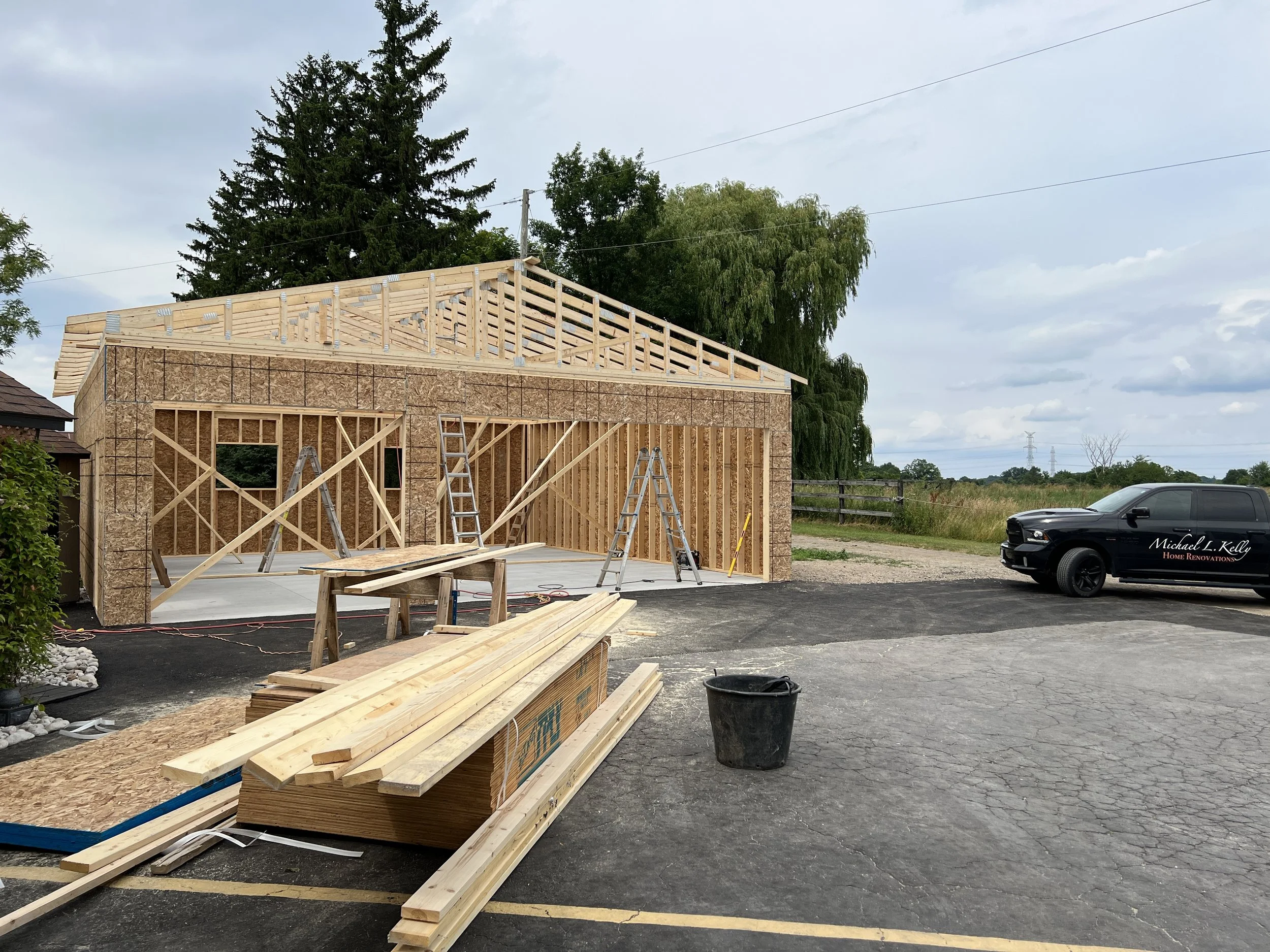 Construction site of a house with wooden framing and OSB sheathing, ladders, and building materials on the ground. A black pickup truck with company branding is parked nearby on a paved area.