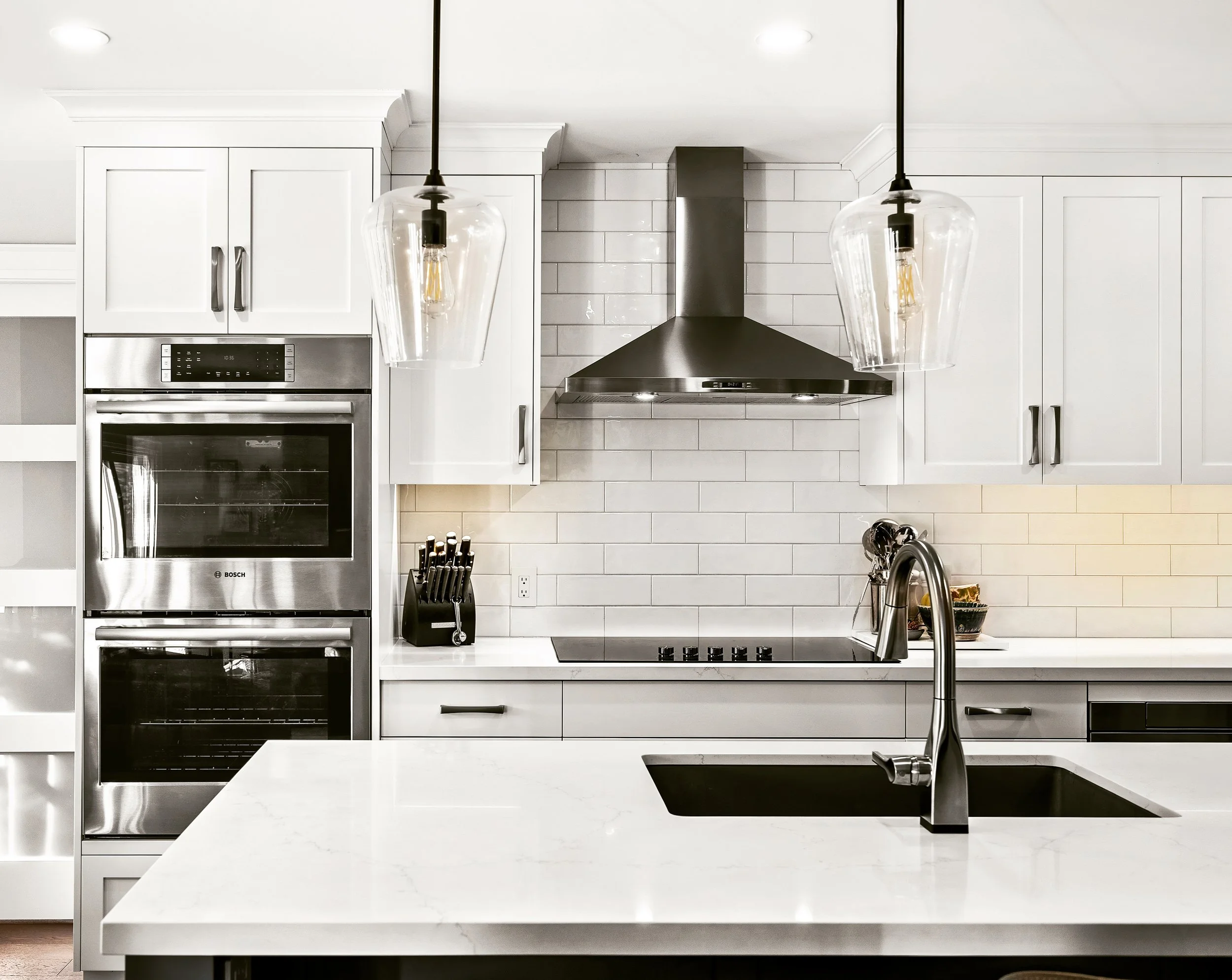 Modern kitchen with white cabinets, a black stovetop, stainless steel oven and range hood, black knife block, and hanging pendant lights.