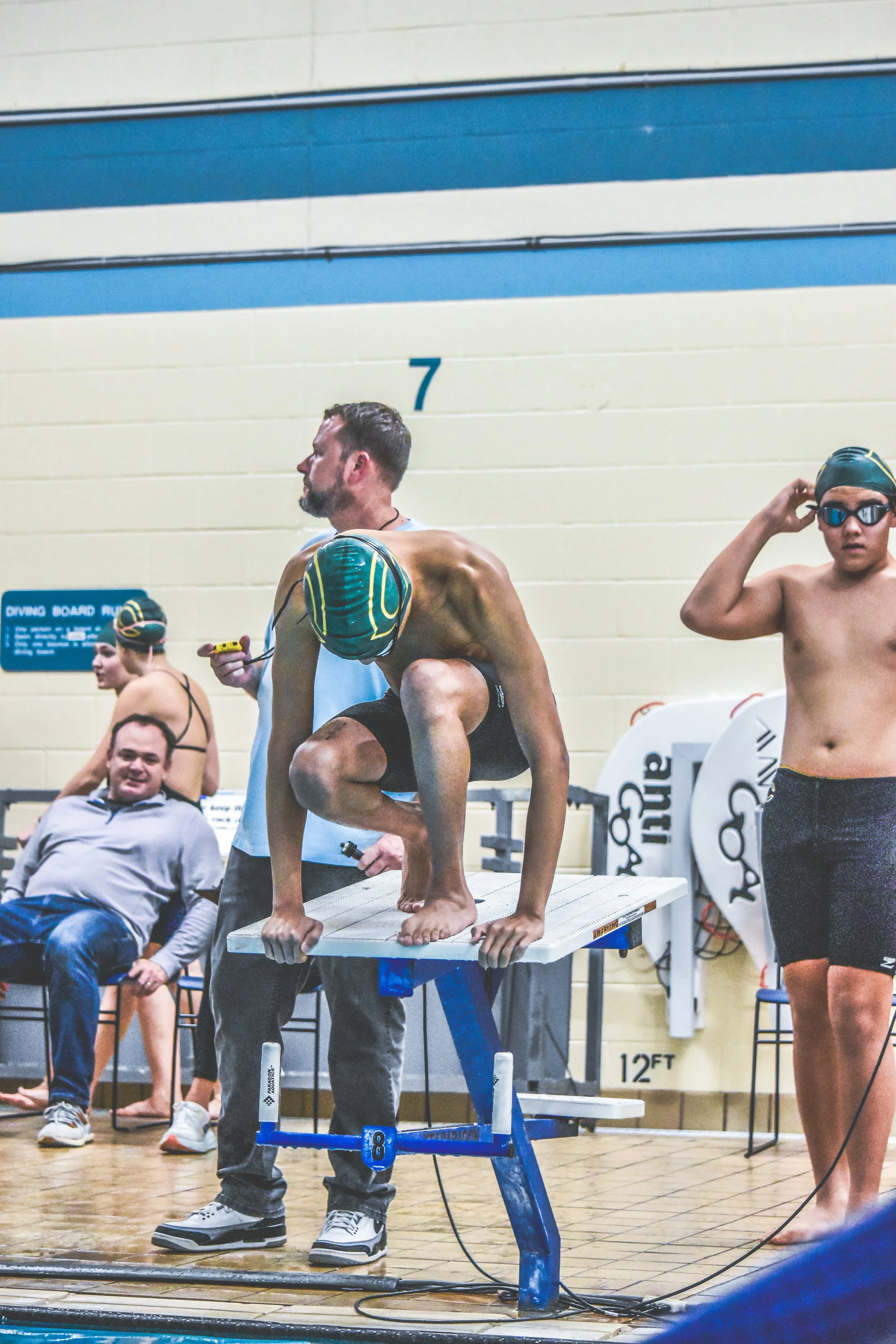 A male swimmer crouches on a starting block at an indoor swimming pool, preparing for a race, with other swimmers and a man seated behind him, and a man adjusting his goggles on the right side.