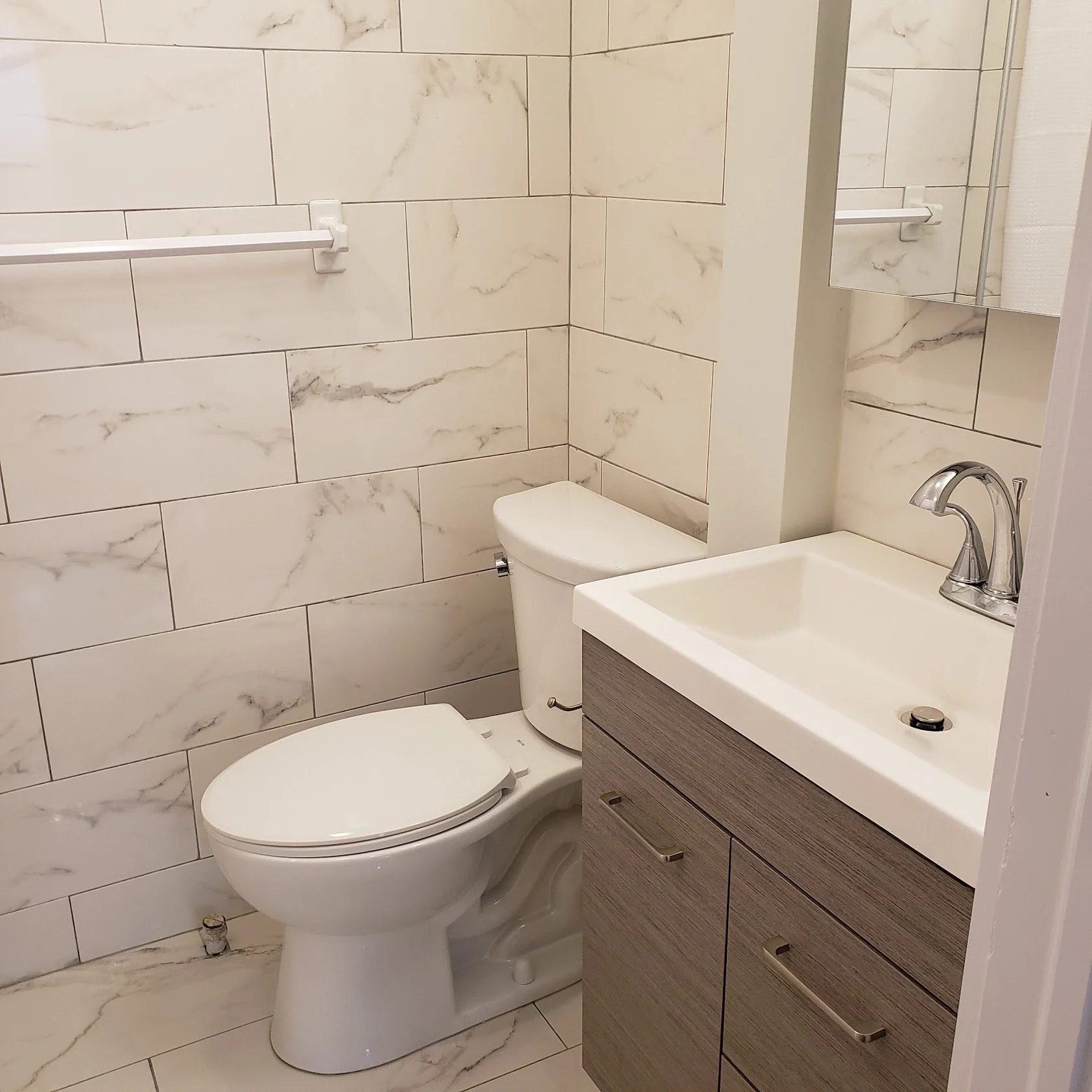 A small bathroom with marble tiled walls, a toilet, a sink with a chrome faucet, and a mirror above the sink.