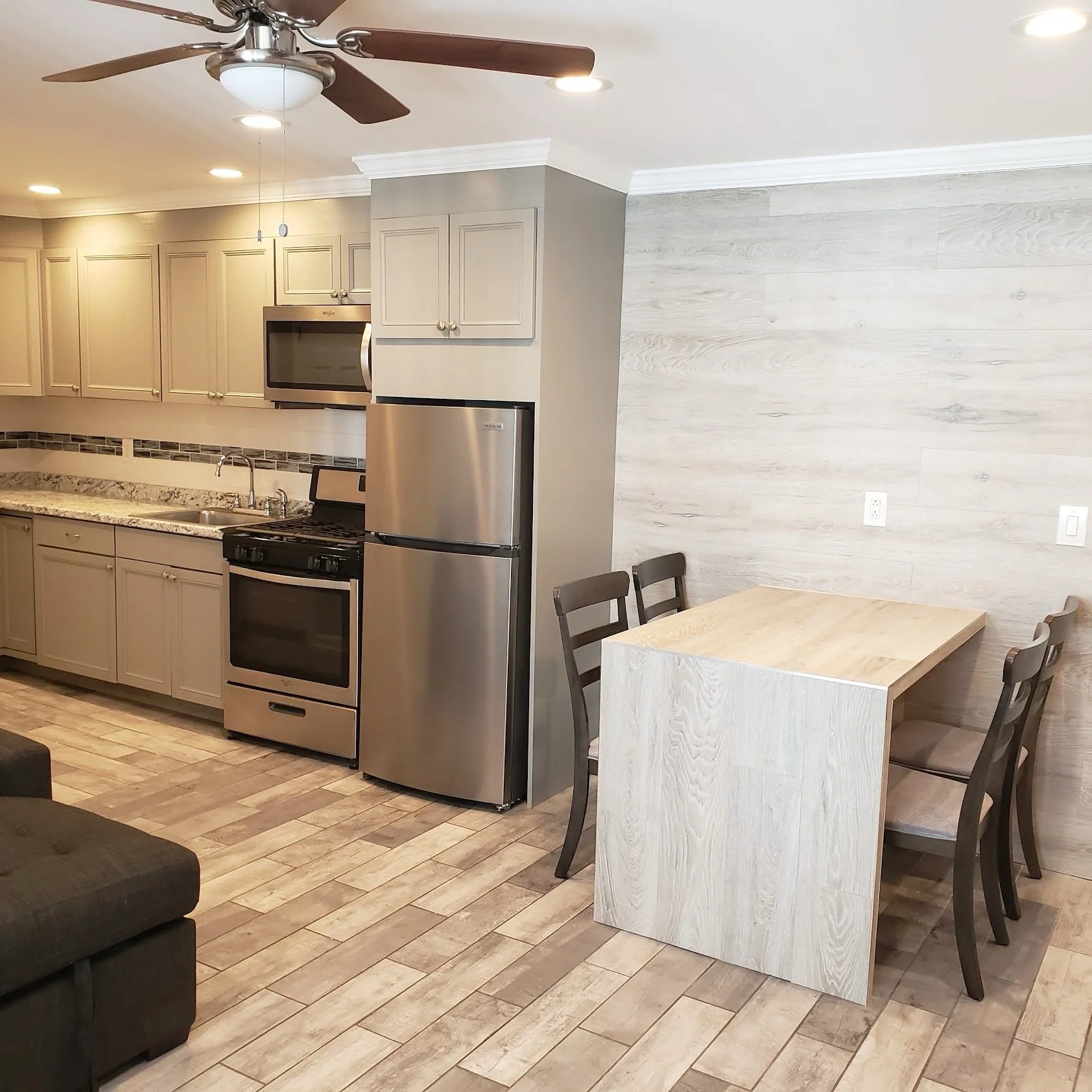 Kitchen with beige cabinets, stainless steel appliances, including a microwave and refrigerator, dark wood ceiling fan, and a dining area with a light wood table and dark wood chairs. The floor is wood-look tile and the wall to the right has textured