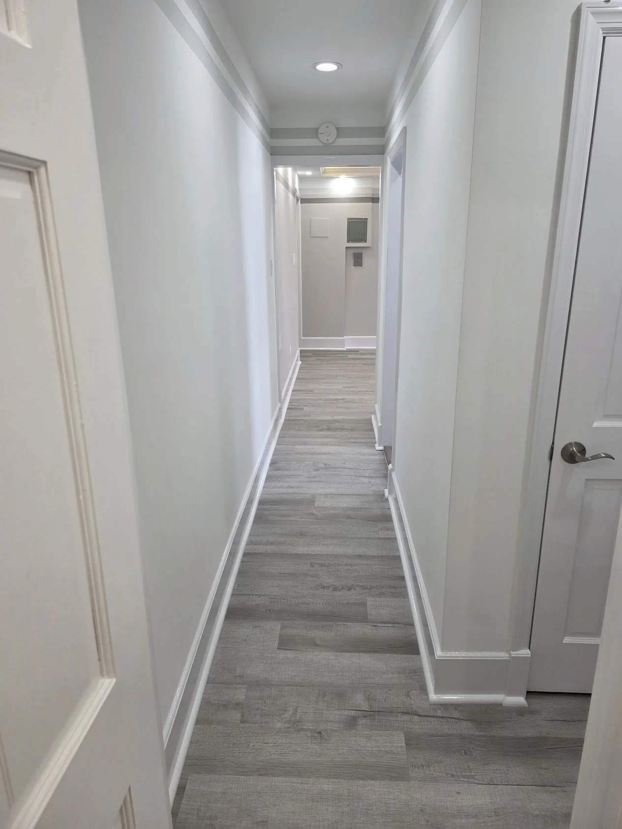 Empty hallway with light gray walls, white trim, wood-style flooring, and doors on both sides.