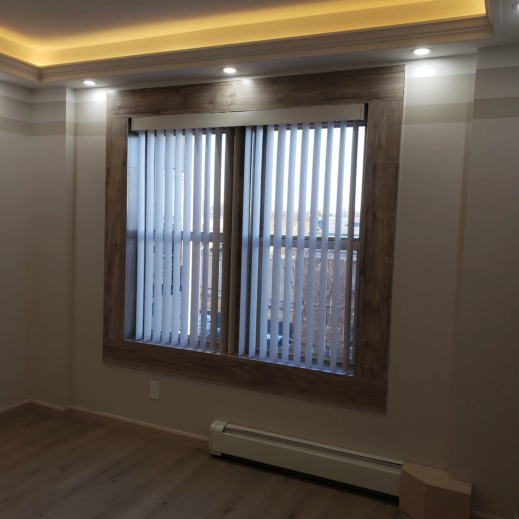 Room with a window framed in wood, vertical blinds covering the window, and ceiling lights with a textured ceiling. The room has light-colored walls and wood flooring.