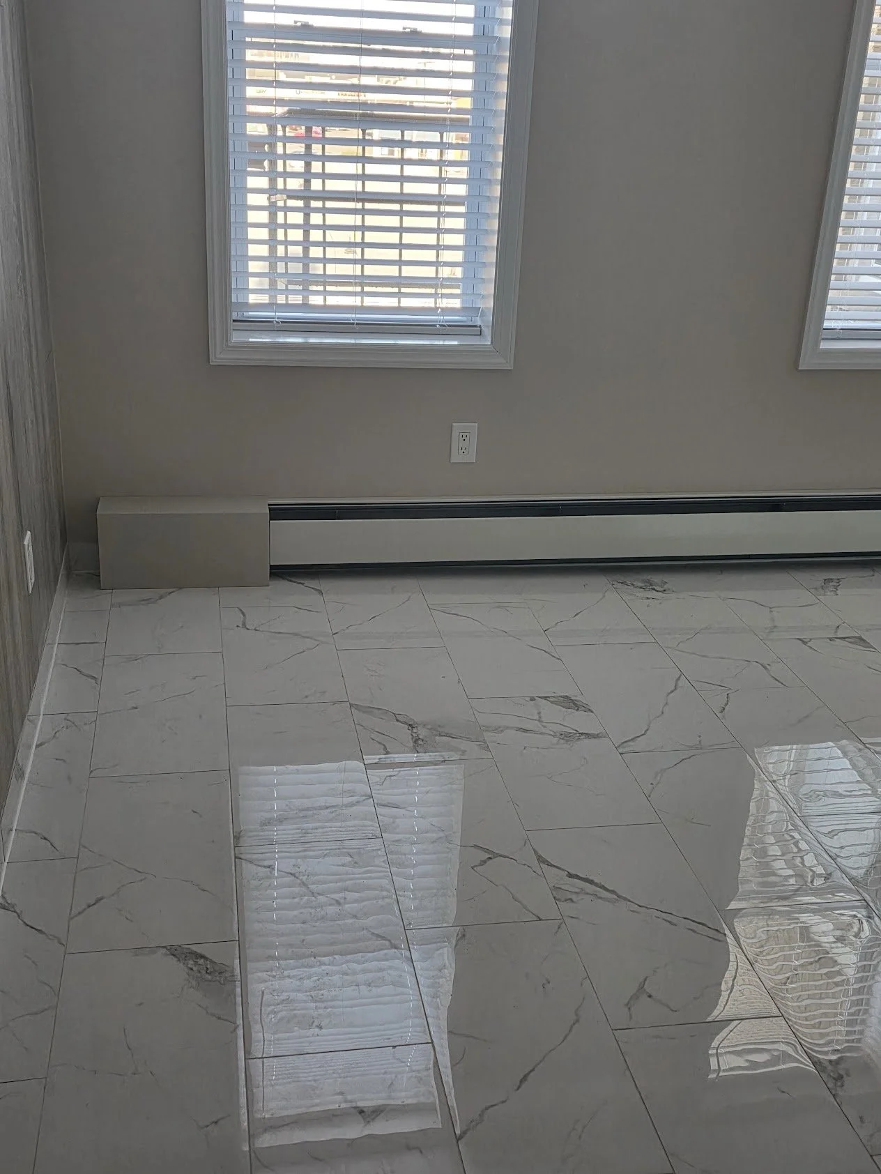 Empty room with two windows and white blinds, white baseboard heater, beige wall, and shiny marble tile floor.