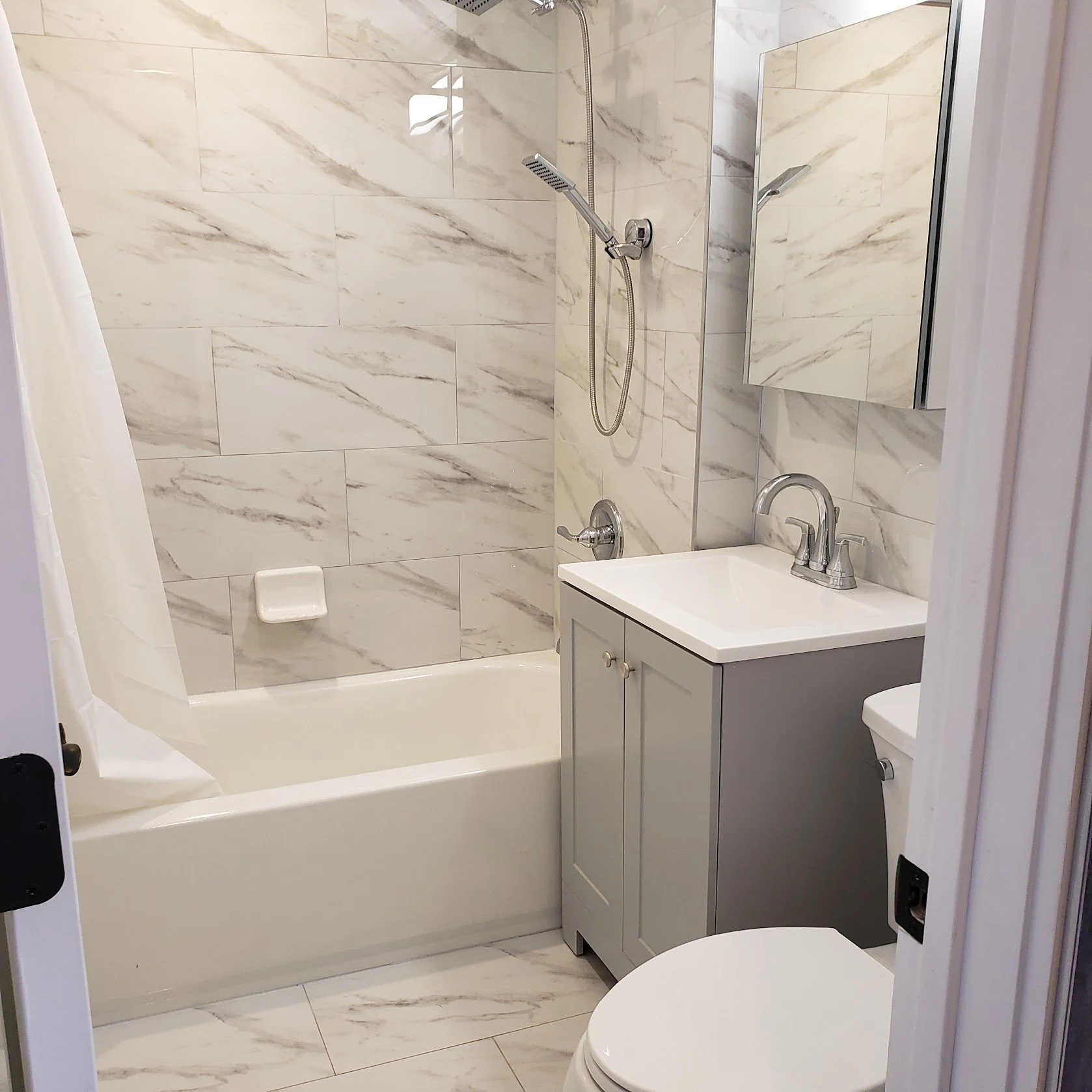 Small bathroom with a shower area, marble tiled walls, a mirror cabinet, a small gray vanity with a sink and faucet, and a toilet.