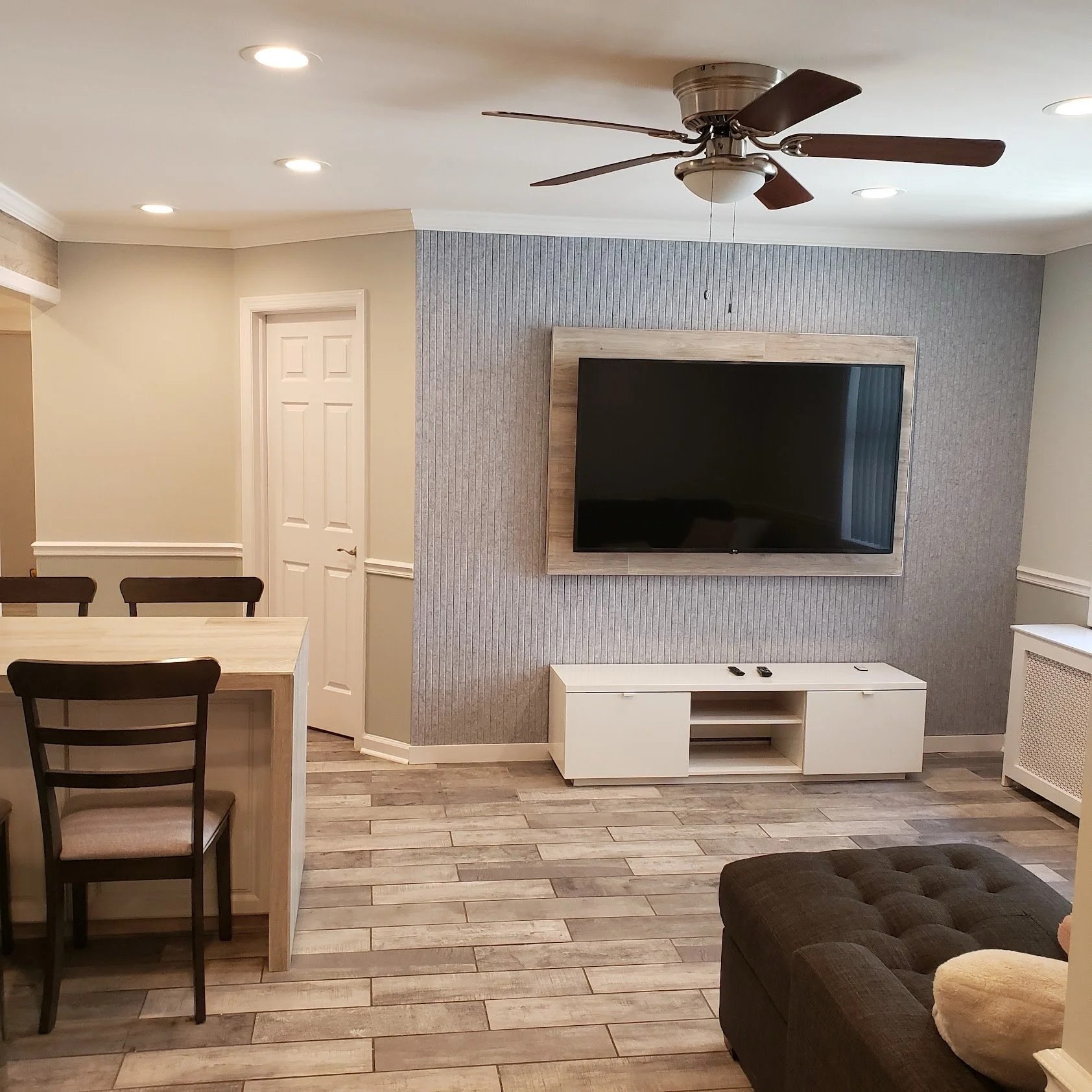 Living room with a wall-mounted flat-screen TV, white entertainment stand, wooden dining table with chairs, textured gray accent wall, ceiling fan, and recessed ceiling lights.