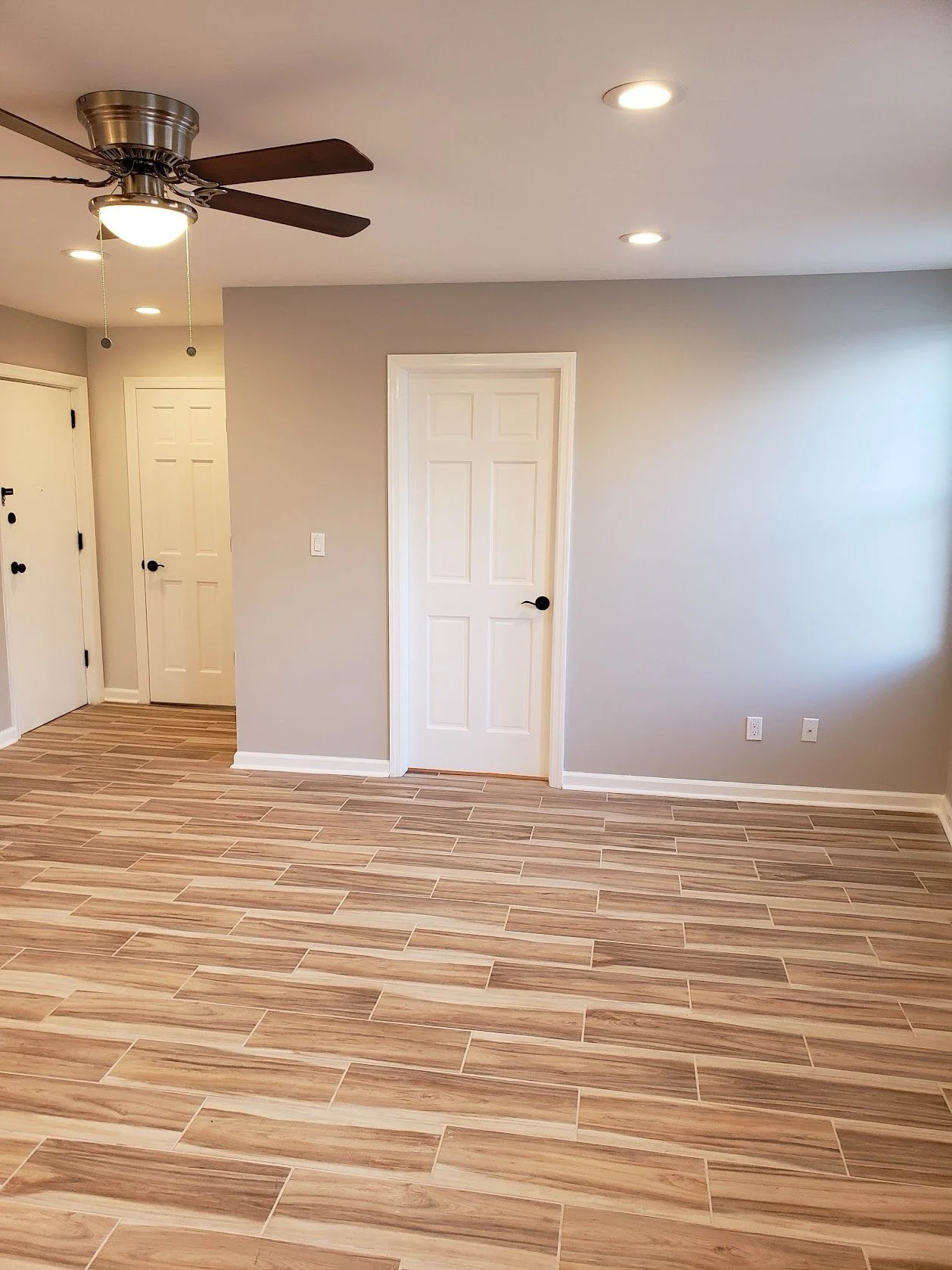 Empty room with wood-look tile flooring, light gray walls, a white door, ceiling fan with dark blades, and recessed lighting.