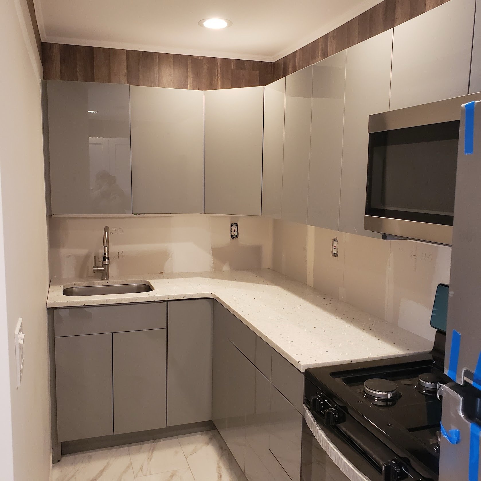 A kitchen with grey cabinets, a black stove with two burners, a white speckled countertop, a stainless steel sink, and an overhead light. There are walls with wood paneling and electrical outlets.