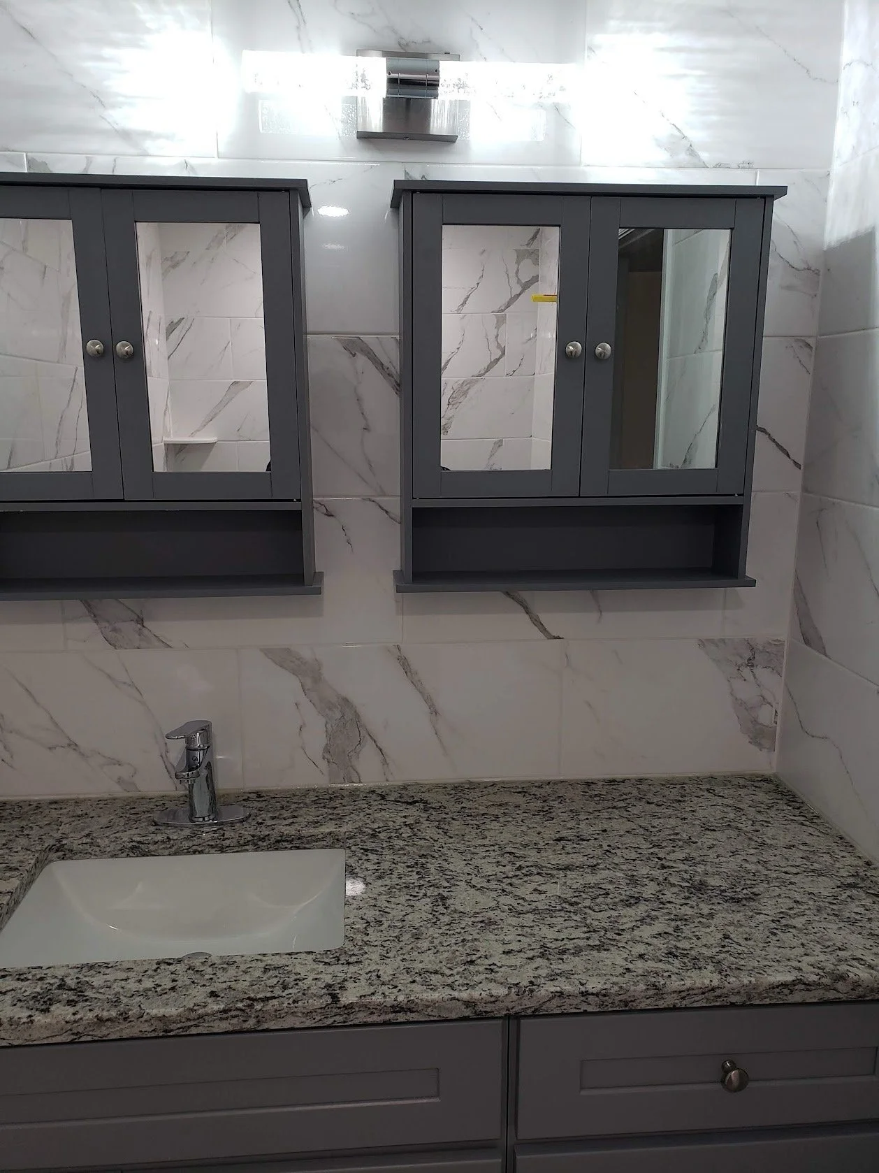 A bathroom with a granite countertop, a white sink, a gray cabinet under the sink, and gray medicine cabinets with glass doors mounted on a marble wall.