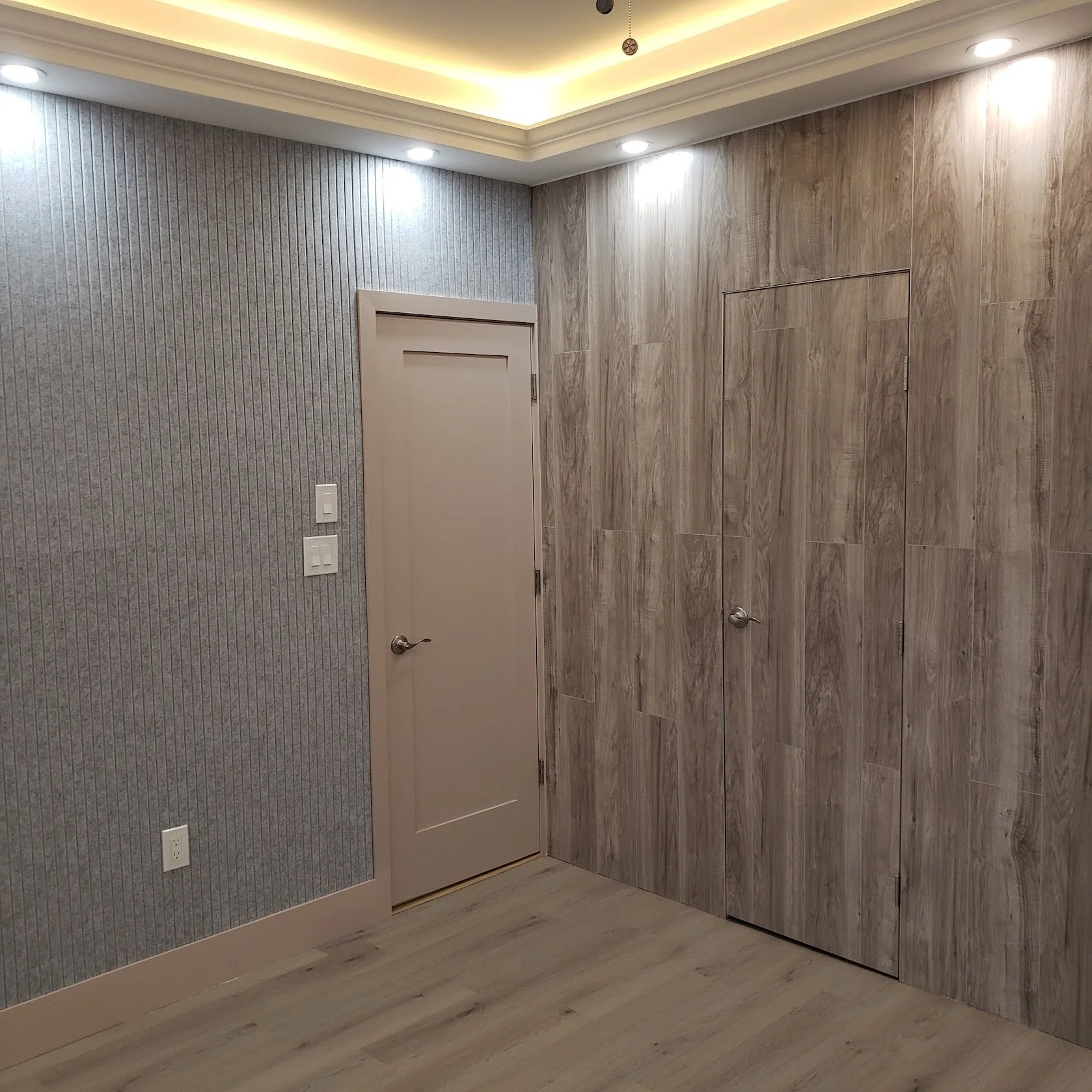 Corner of a room with textured gray and wood-paneled walls, ceiling with recessed lighting, and a closed door with a door handle.